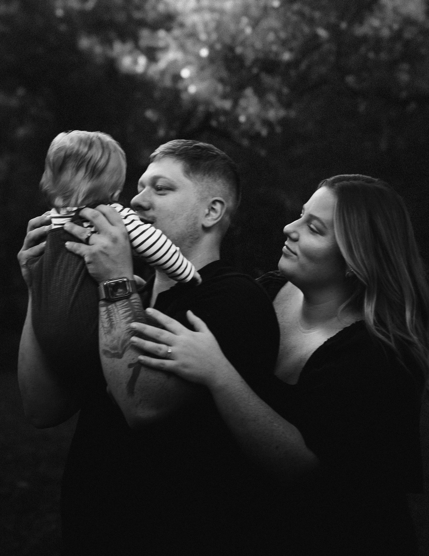 A black and white portrait of a loving young family with a small child in a casual outdoor setting.