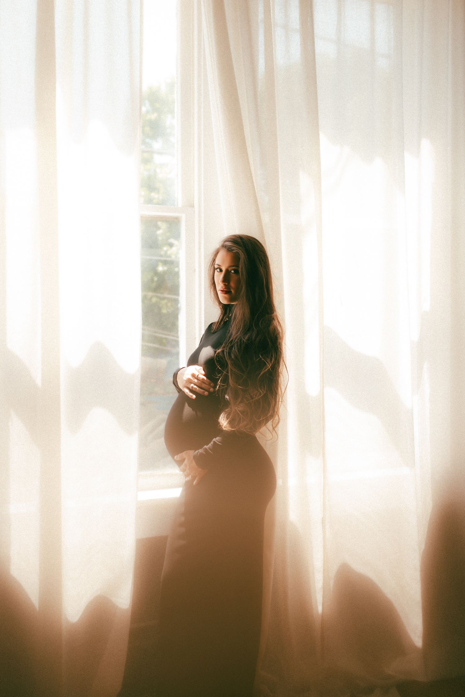 A silhouetted figure stands by sheer curtains in soft natural light creating a dreamy atmospheric portrait.