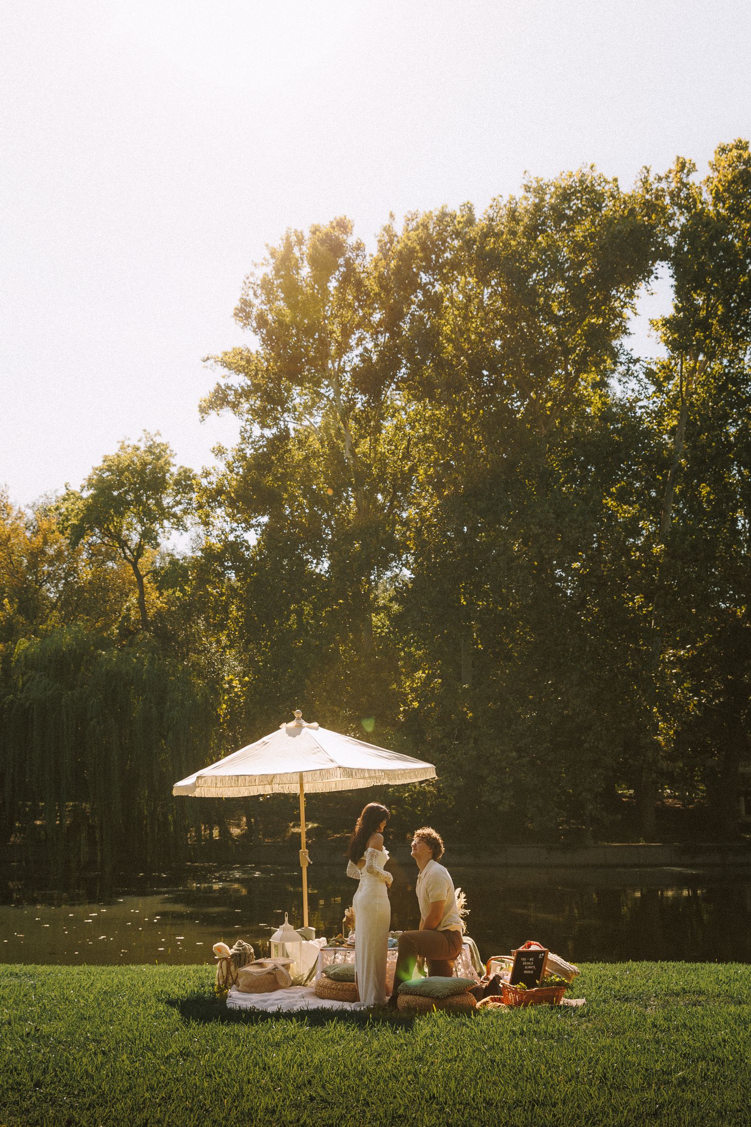 Couple enjoying a romantic sunset picnic on a grassy hill with a white umbrella and trees in the background.