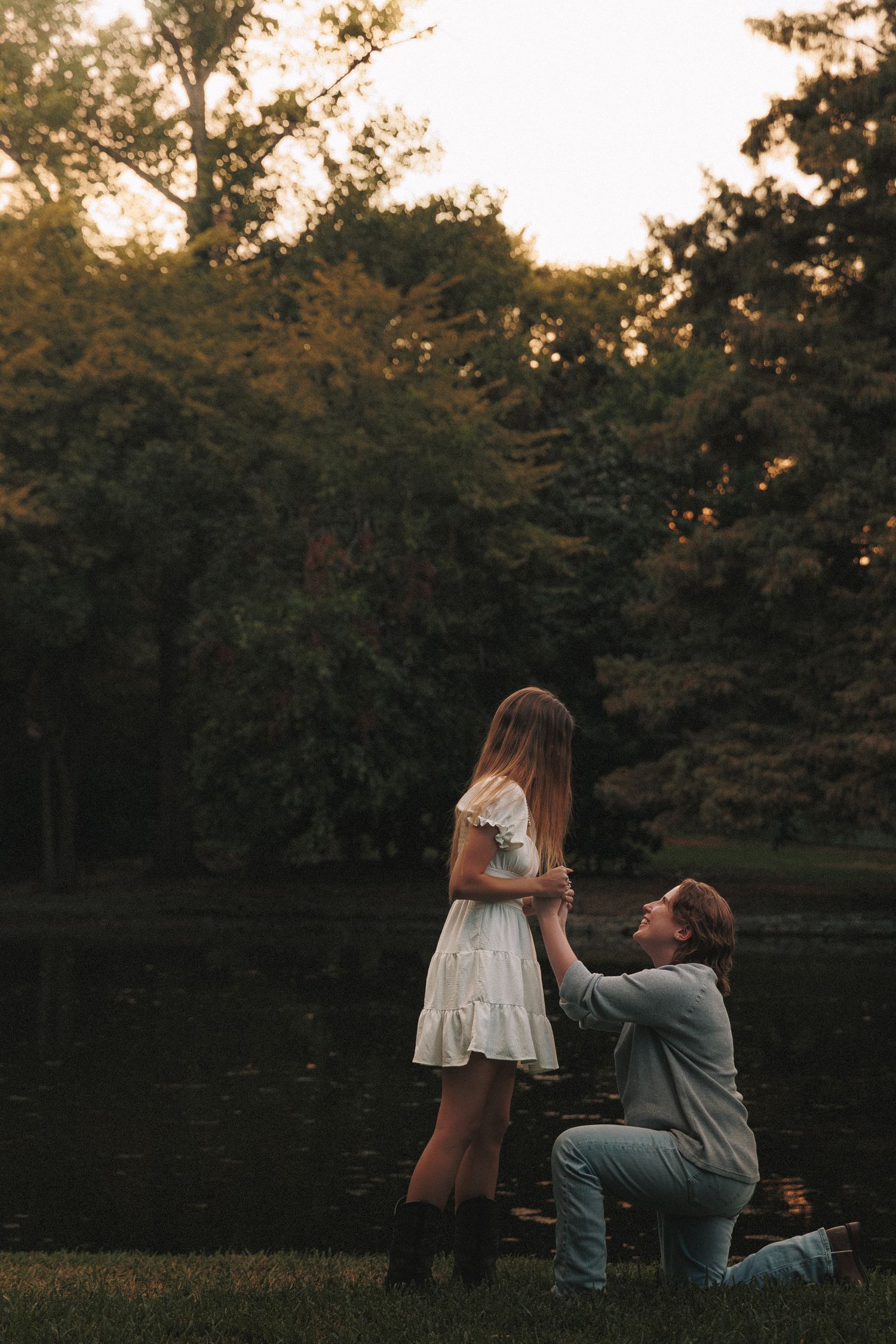A couple shares an intimate moment during sunset by a lake in a wooded area.