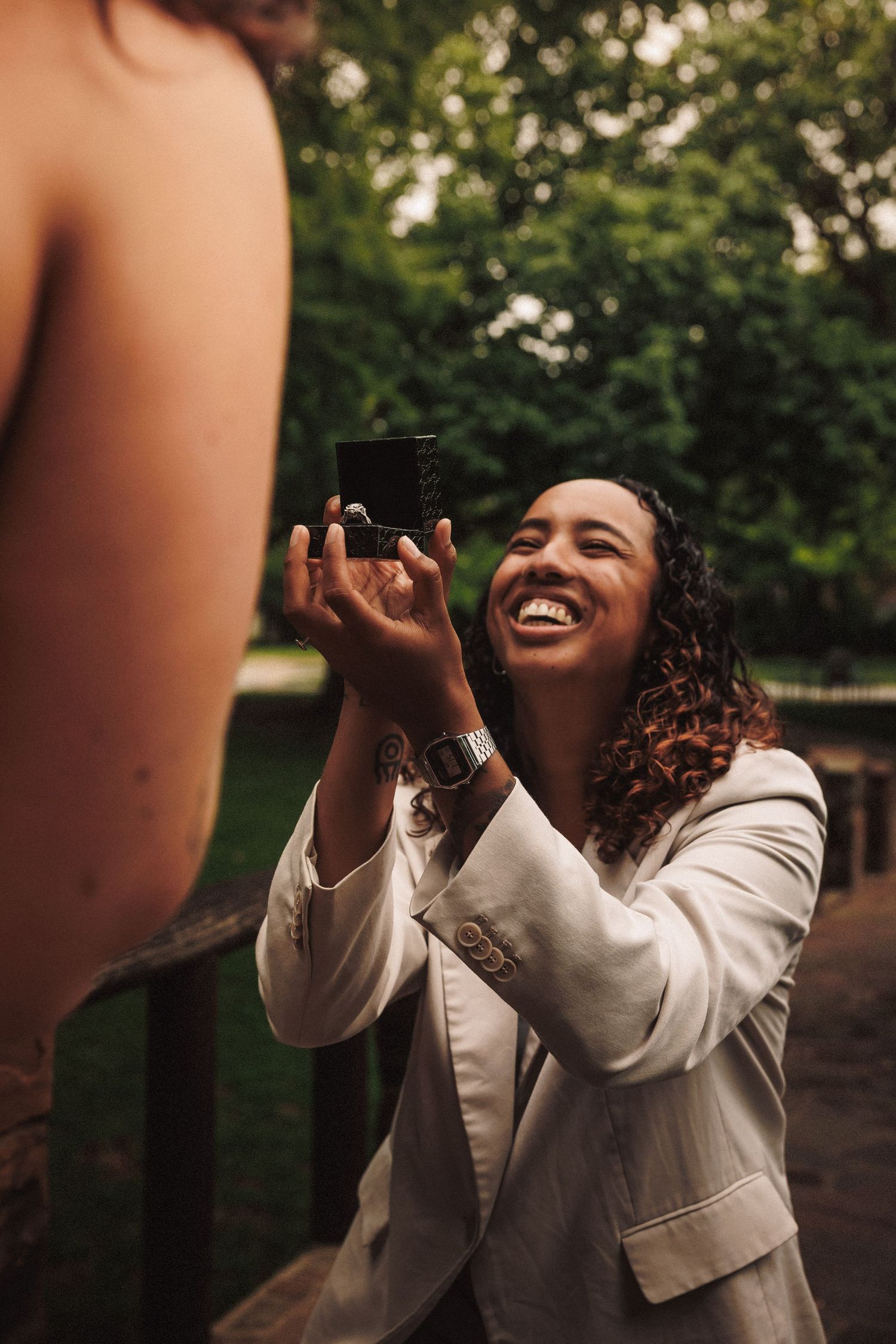 Woman in white jacket laughs joyfully while reaching up with arms extended in an outdoor setting.