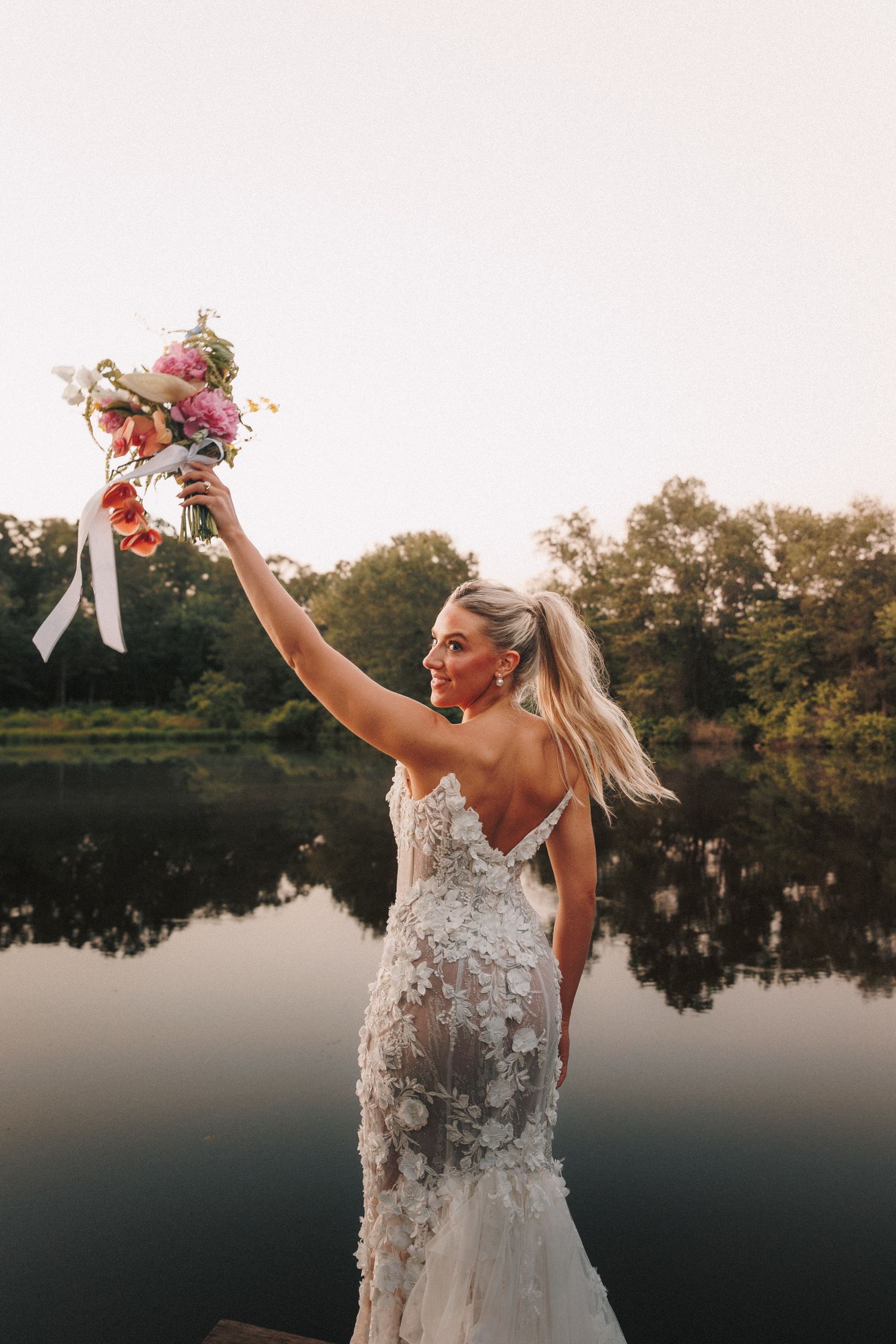 A bride in a floral lace dress celebrates joyfully by a lake at sunset.