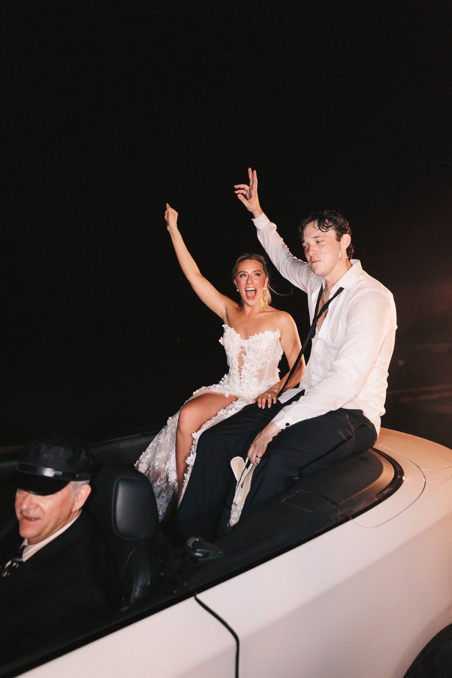 Newlyweds celebrating while riding in the back of a white car at night.