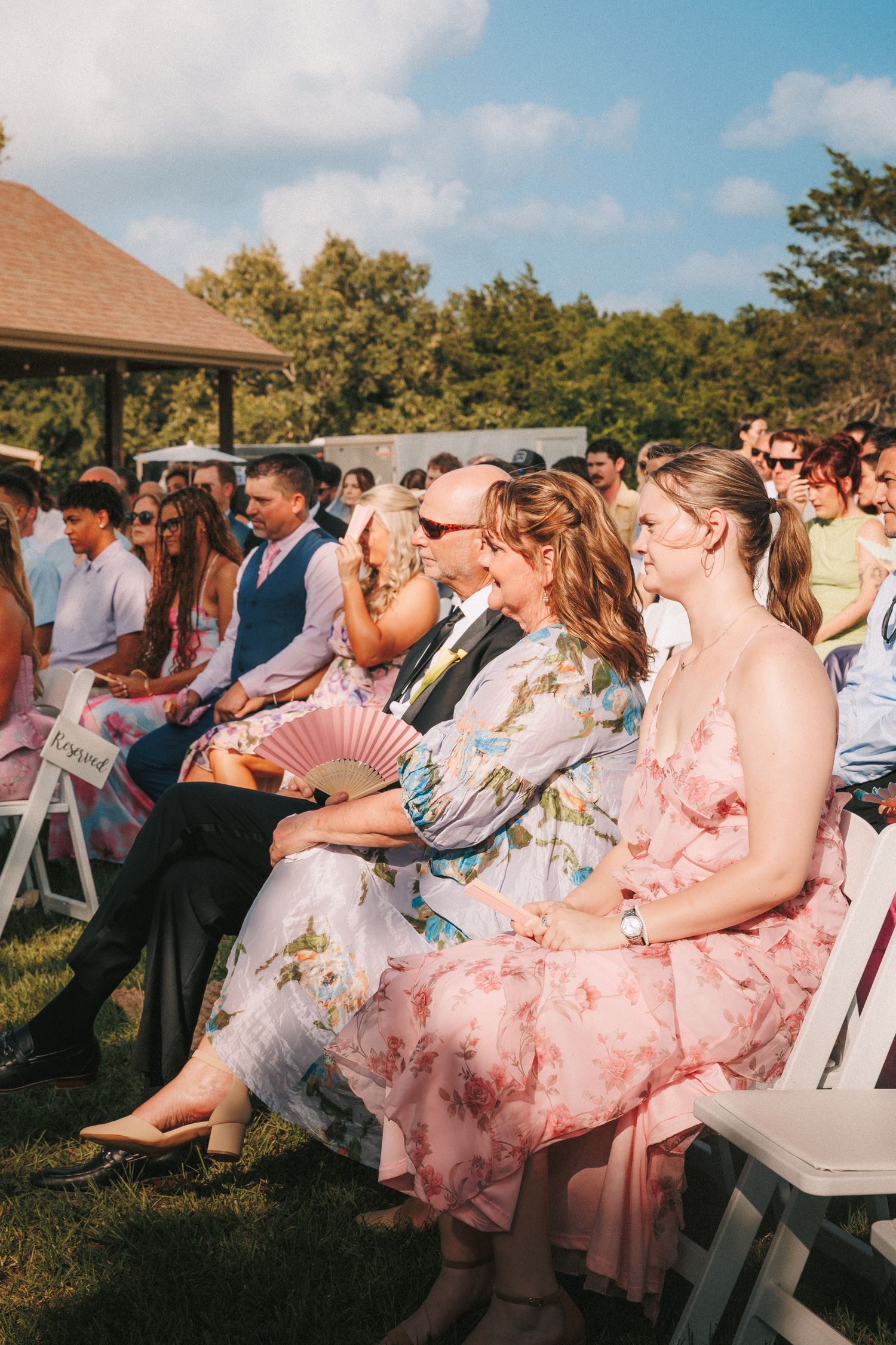 Wedding guests seated in white chairs wearing pastel and floral dresses during an outdoor ceremony.