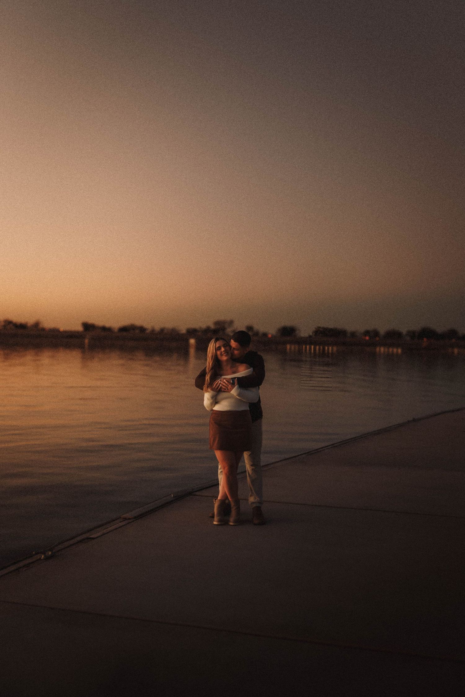 A distant view of a couple sharing a romantic moment on a dock at dusk.