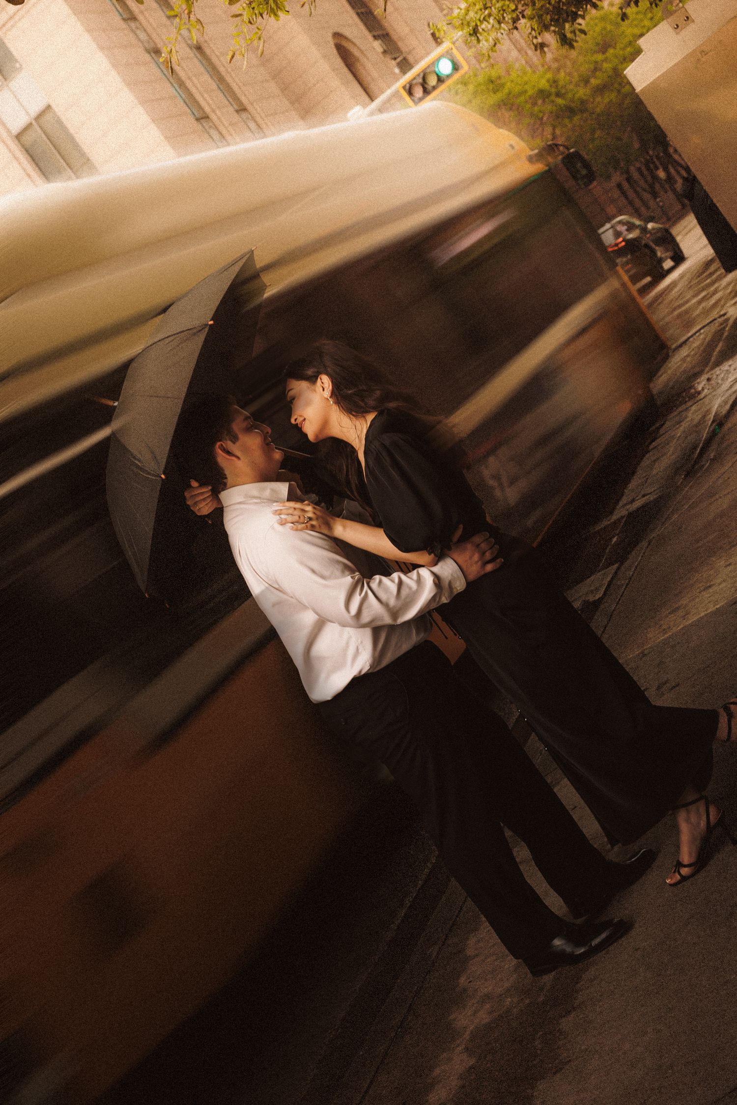 Couple sharing a romantic kiss under an umbrella on a city street at night, with motion blur from passing traffic.