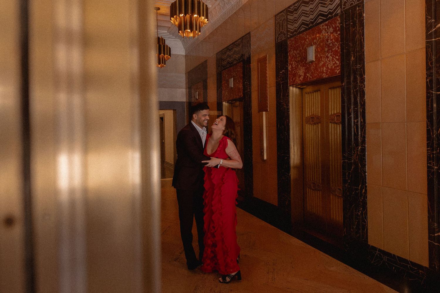 Couple in elegant attire sharing a romantic moment in an Art Deco hallway with ornate marble walls and golden lighting.