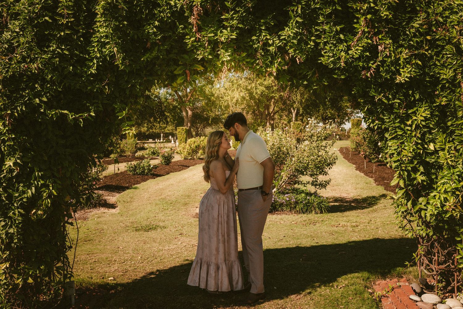 A couple strolls through a lush garden landscape with golden sunlight filtering through the trees.