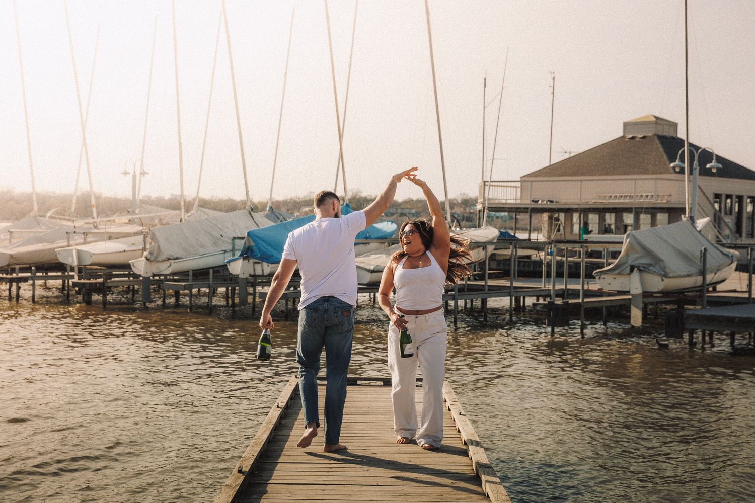 People walking and fishing along a wooden pier at sunset with boats and docks visible in the background.