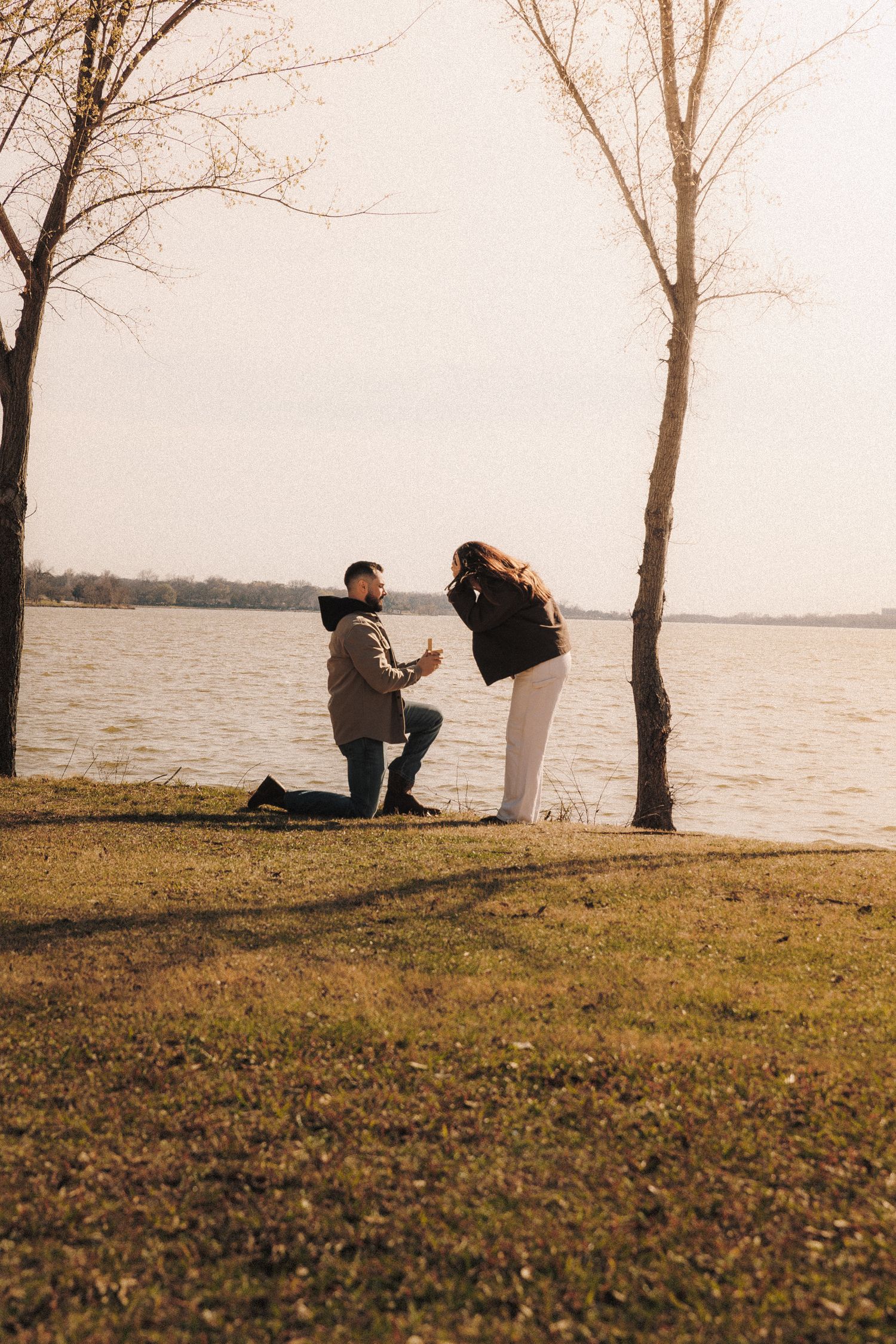 Silhouettes of a couple during a sunset proposal by a lake with bare trees in the background.