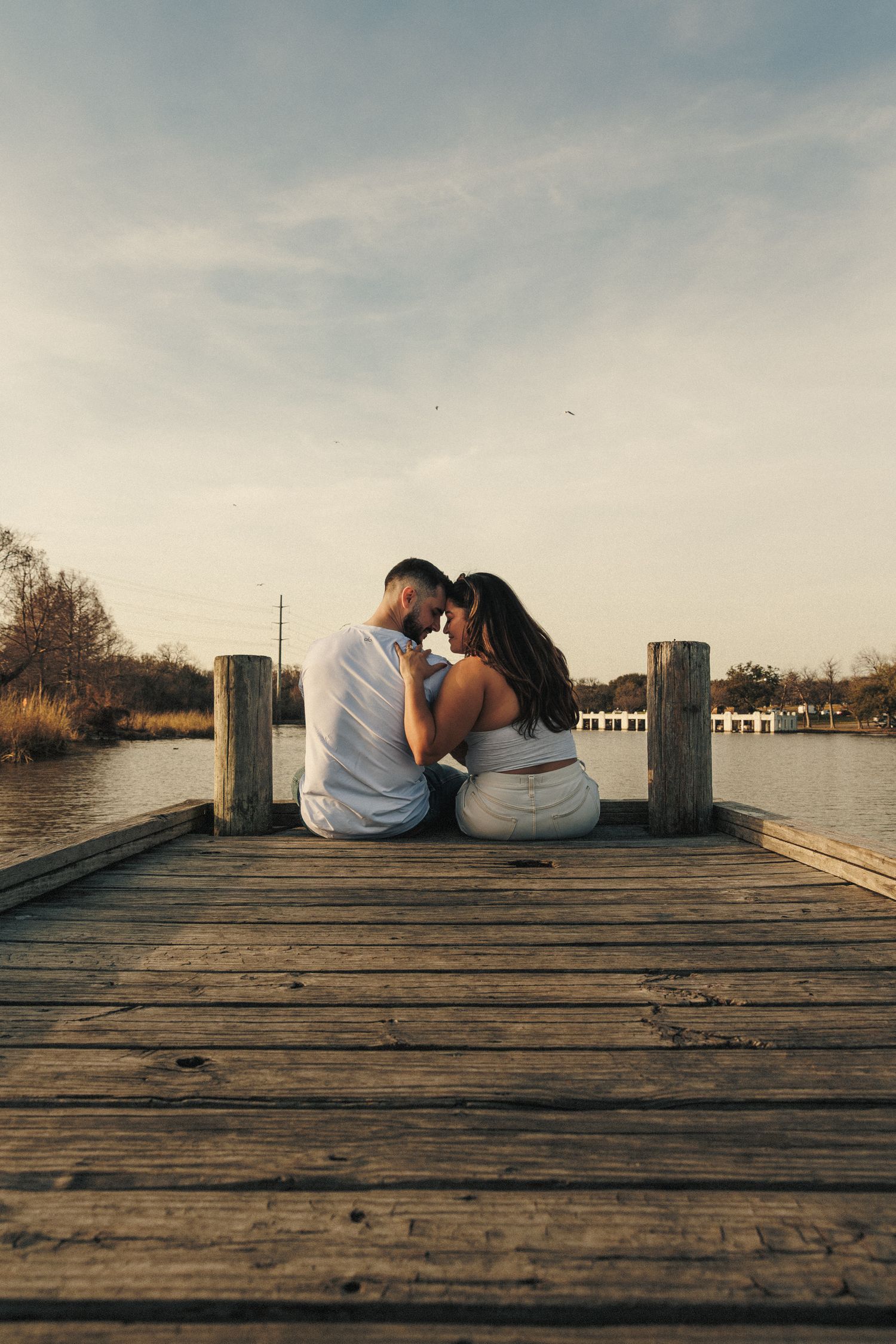 A couple sits close together on a wooden dock overlooking the water.