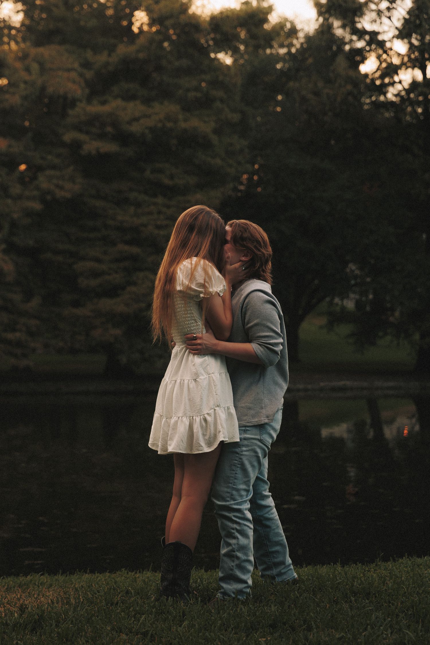 A tender moment between a couple by a lake at sunset, with one wearing a white dress and the other in casual clothes.