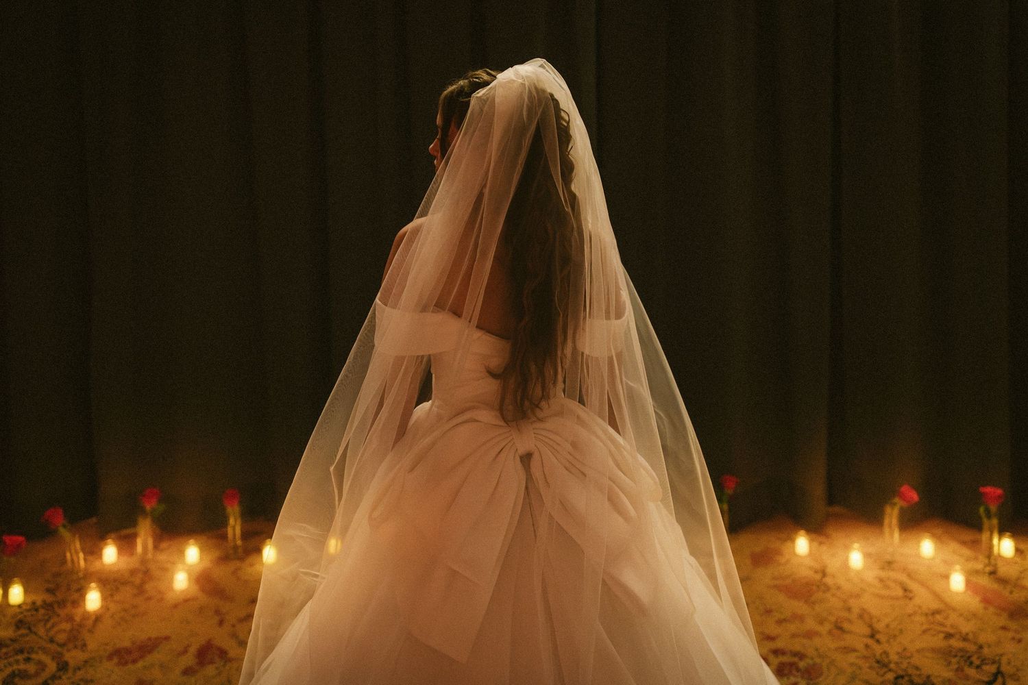 Bride in white ball gown with veil seated among glowing candles in a dimly lit room.