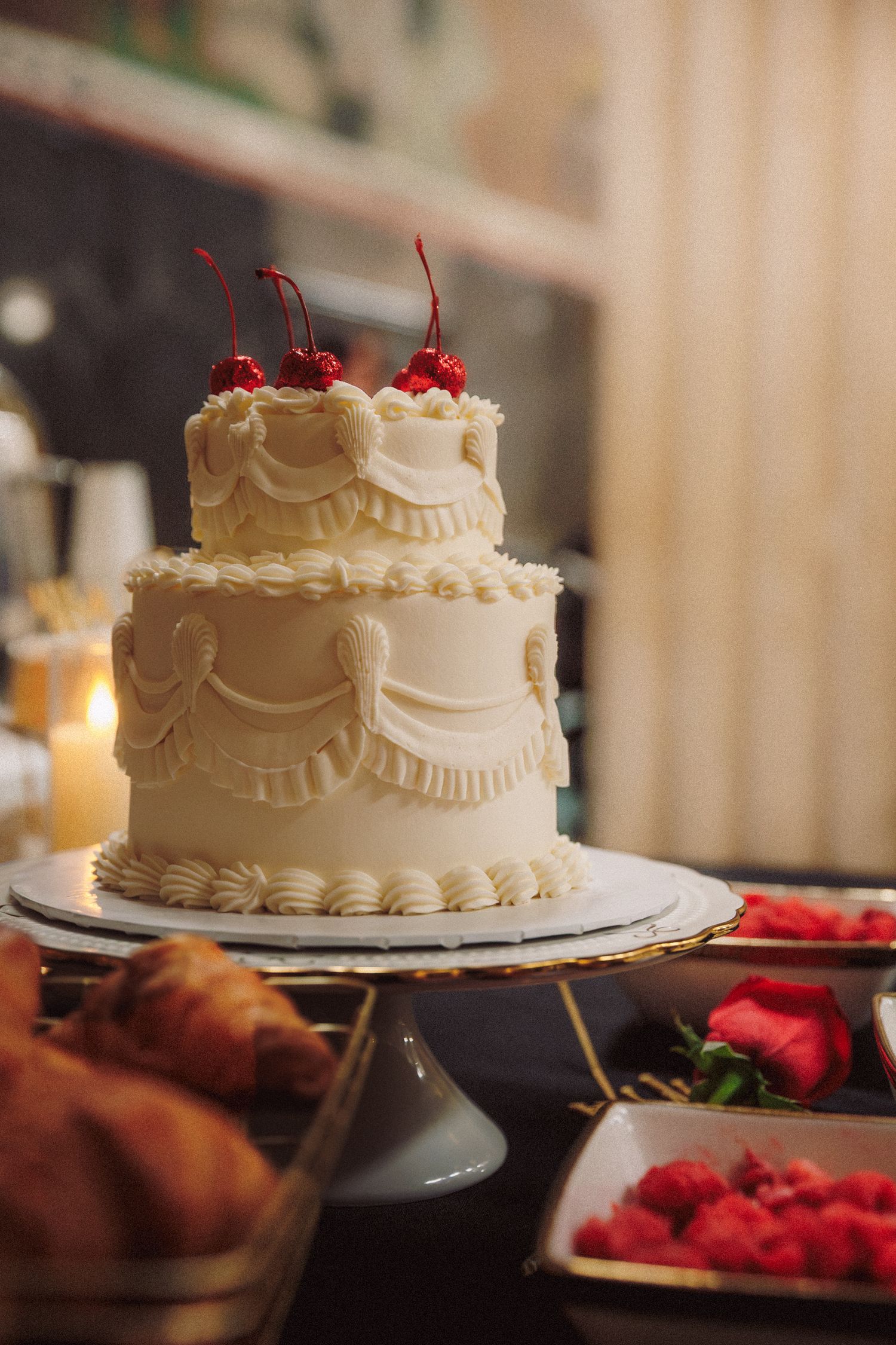 Elegant two-tier white frosted cake topped with red cherries on a cake stand, surrounded by pastries and fresh raspberries.