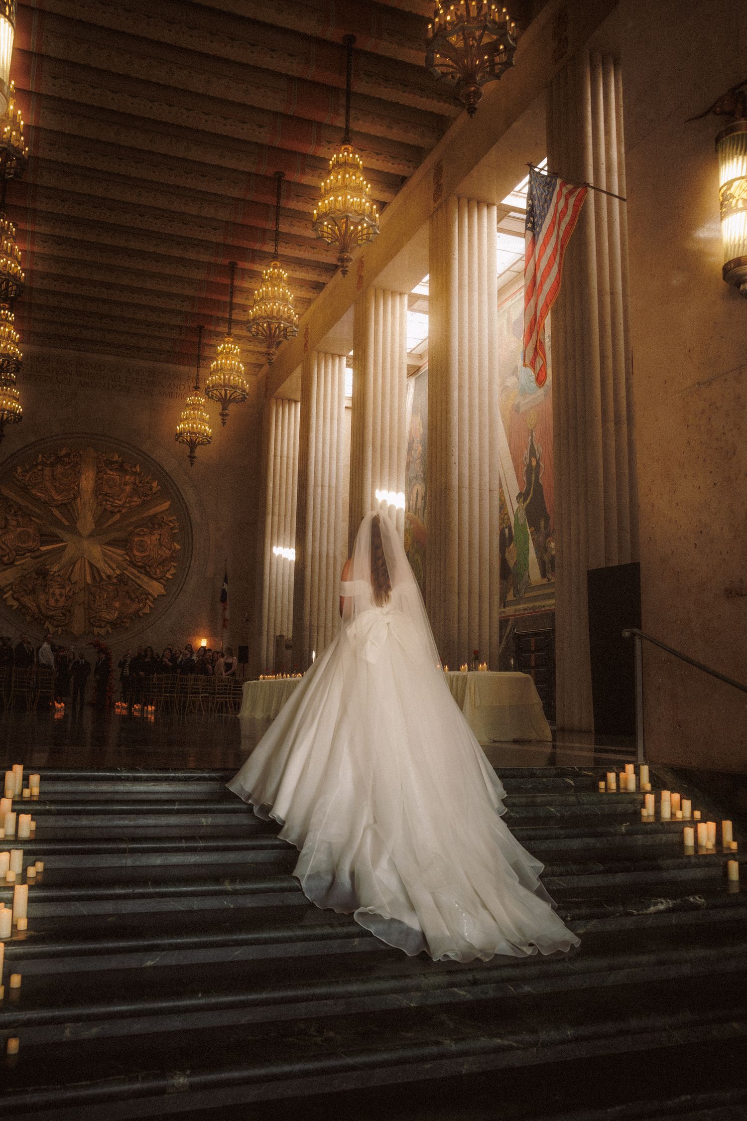 Bride in white gown with long veil walking up candlelit steps in grand hall with columns.