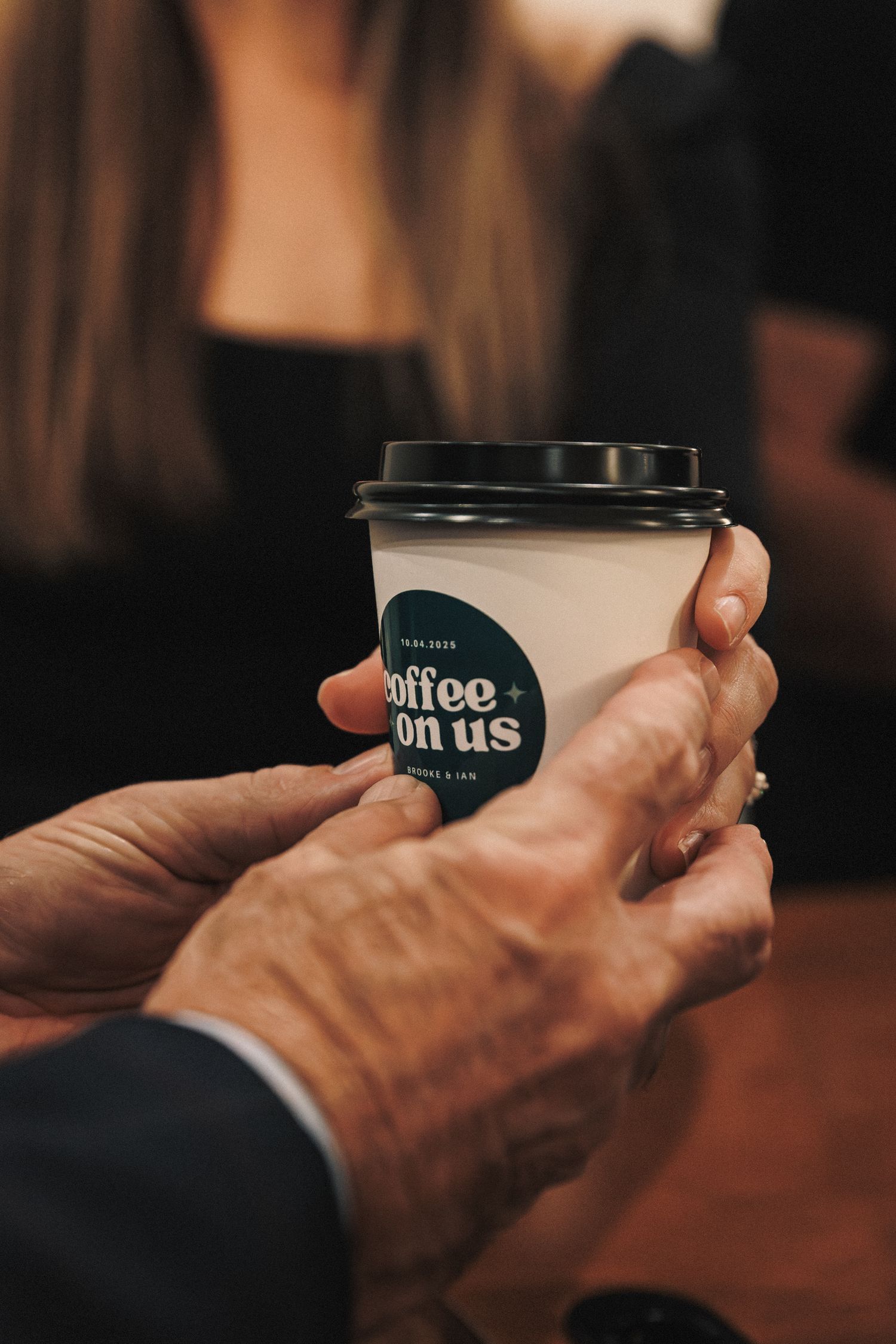 Hands holding a branded coffee cup with coffee on us sticker at a wedding reception event.