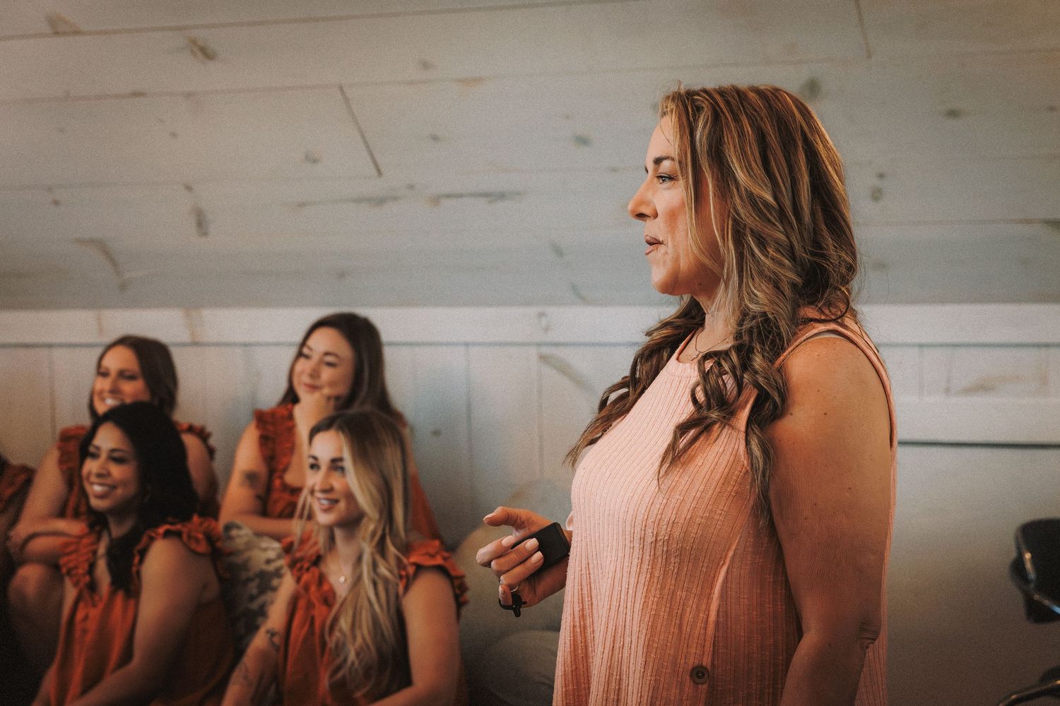 Bridesmaid in blush pink dress speaking to seated bridesmaids in warm rustic venue.