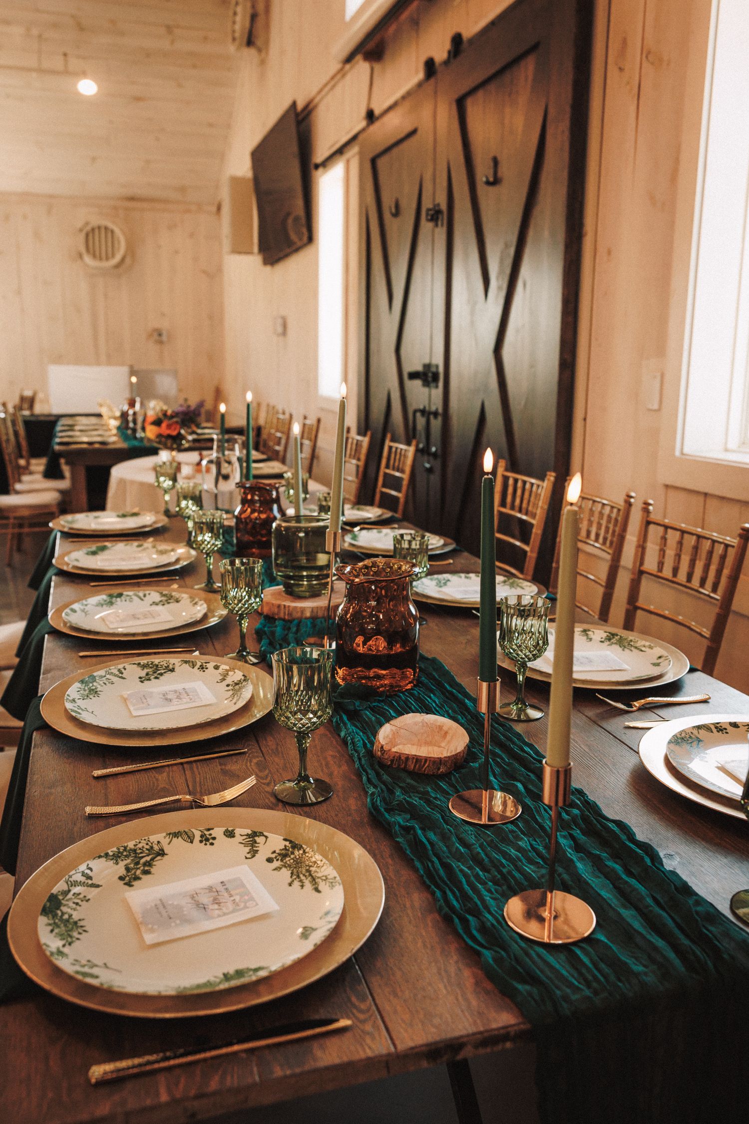 Elegant rustic barn wedding reception table with teal runner, gold chargers, floral plates, taper candles, and chiavari chairs.