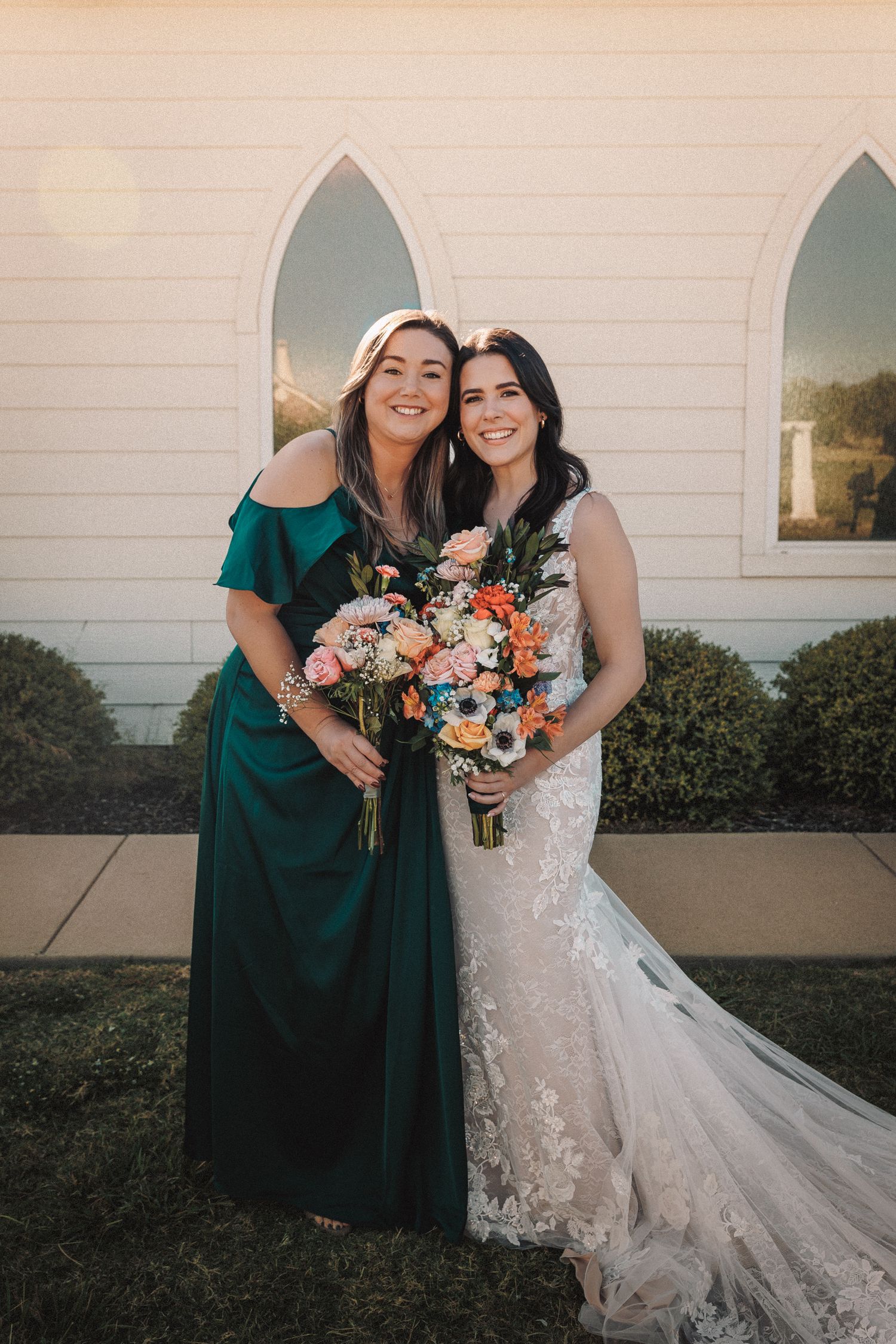 Bride in lace gown holds colorful bouquet with bridesmaid in emerald dress outside white chapel.