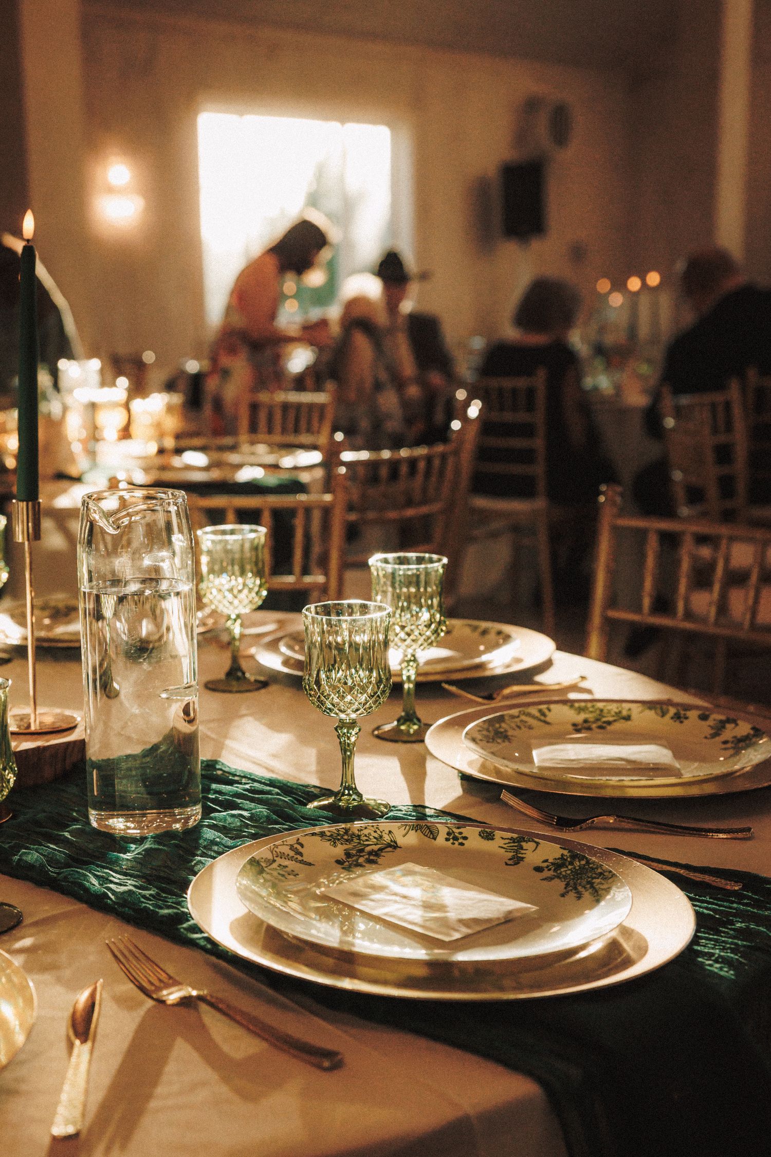 Elegant wedding reception table set with green stemware, floral plates, gold cutlery, and dark green runner in warm candlelit venue.