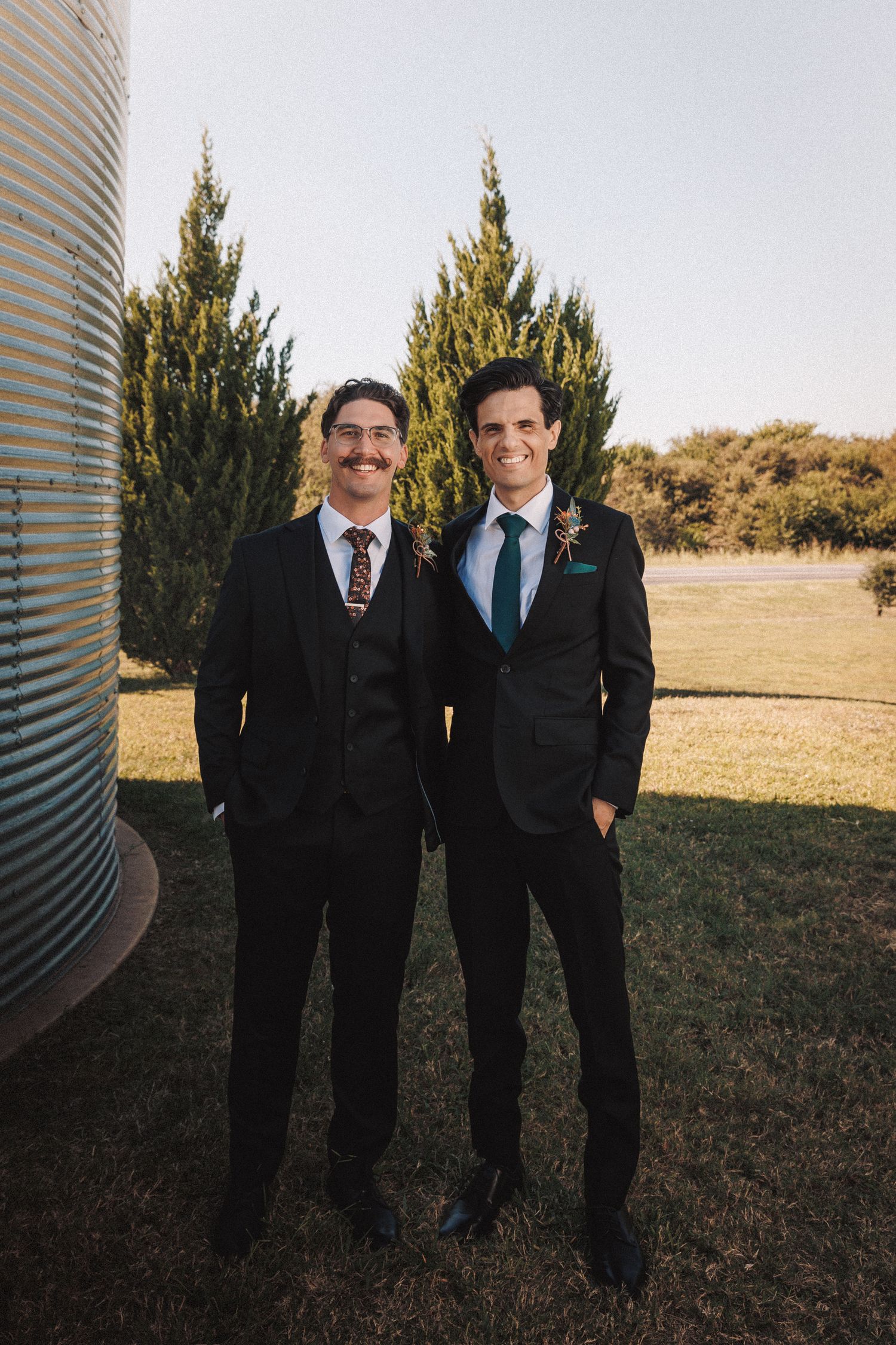 Two groomsmen in black suits smiling together outdoors near grain silo at wedding.