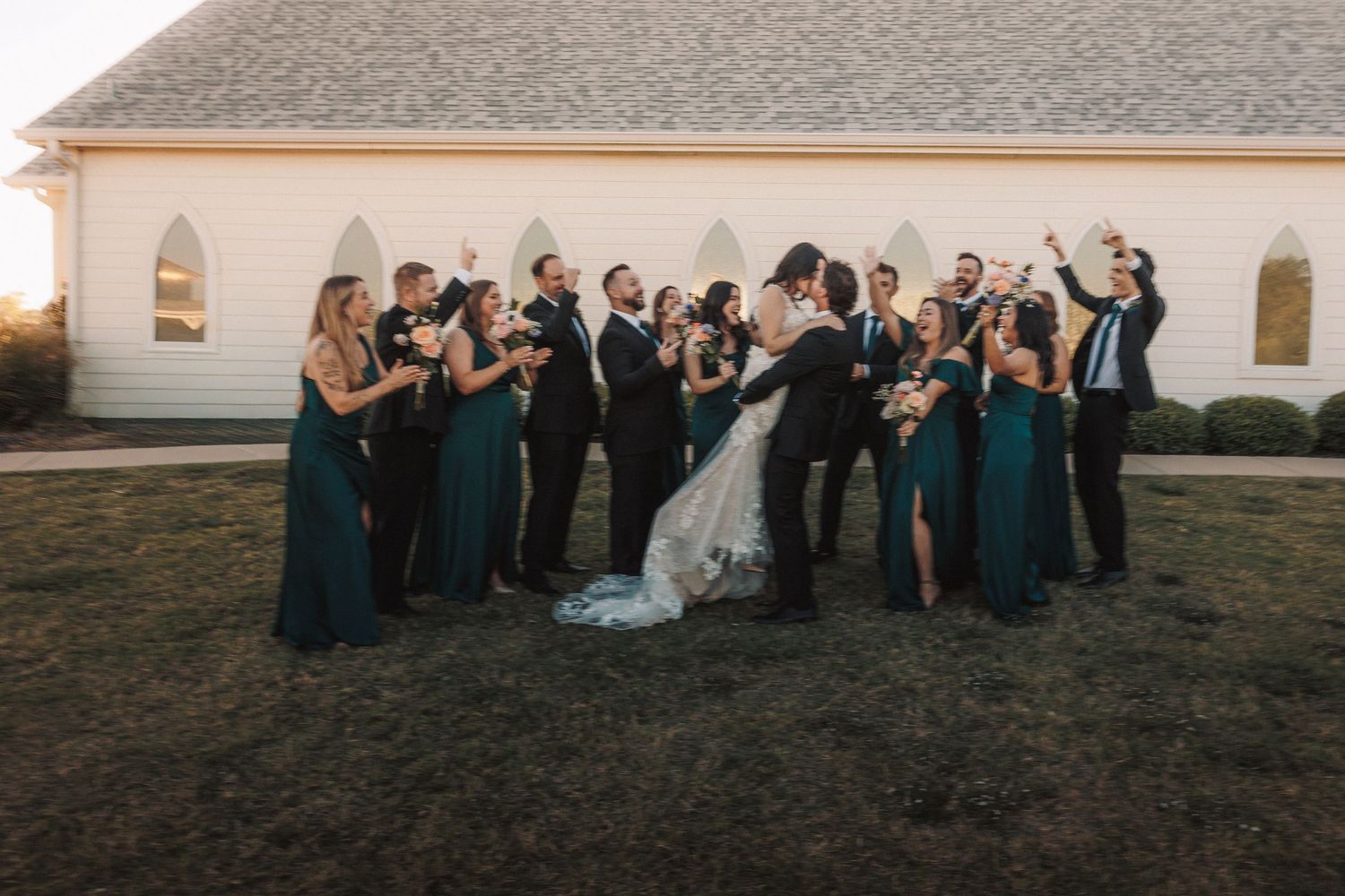 Wedding party in teal dresses and black suits celebrating outside white chapel at golden hour.