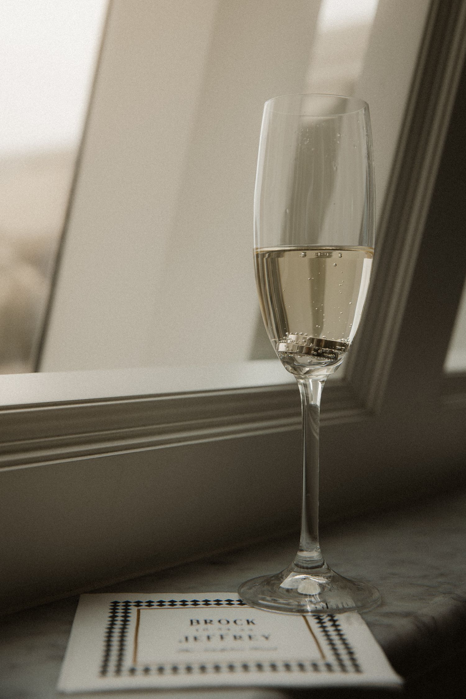 Champagne flute filled with sparkling wine beside a wedding invitation card reading Brock and Jeffrey on a windowsill.