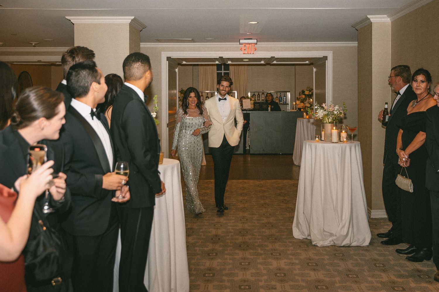 Groom in ivory tuxedo makes grand entrance at wedding reception as guests mingle with champagne.