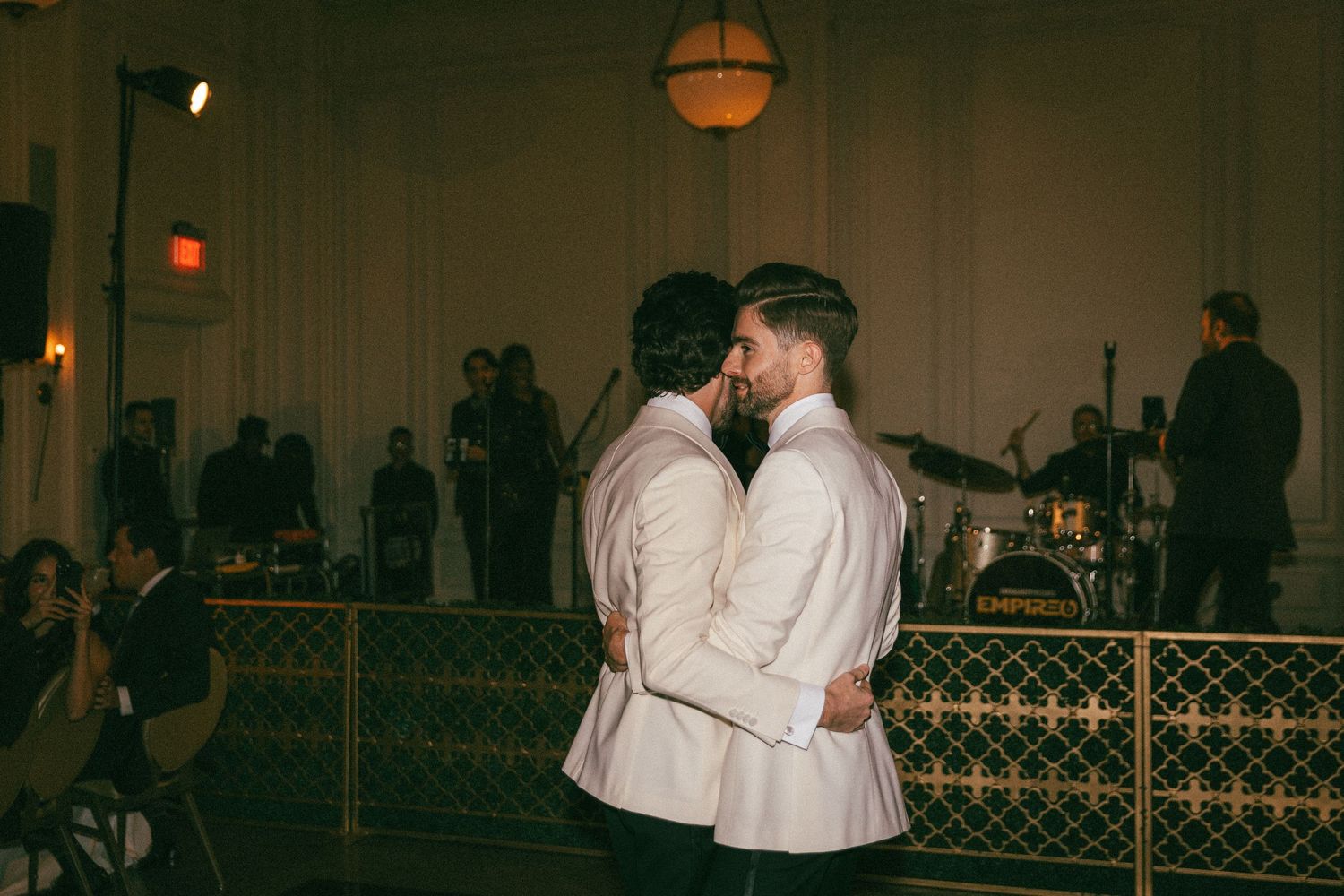 Two grooms sharing first dance kiss in elegant ballroom with live band backdrop.