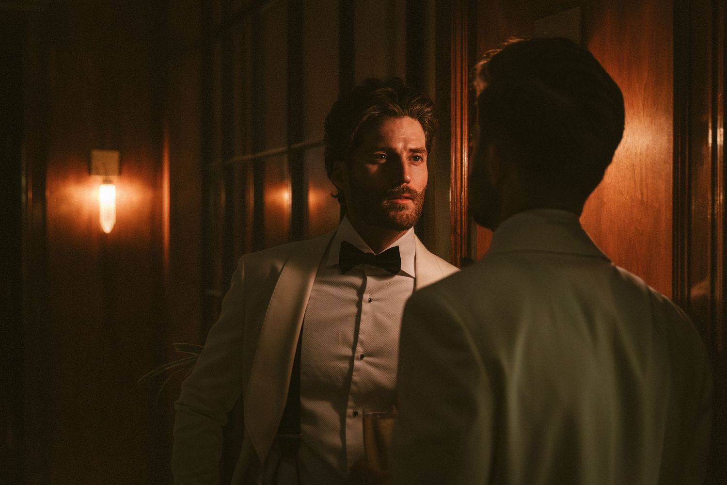Groom in white tuxedo with bow tie standing in elegant wood-paneled hallway.