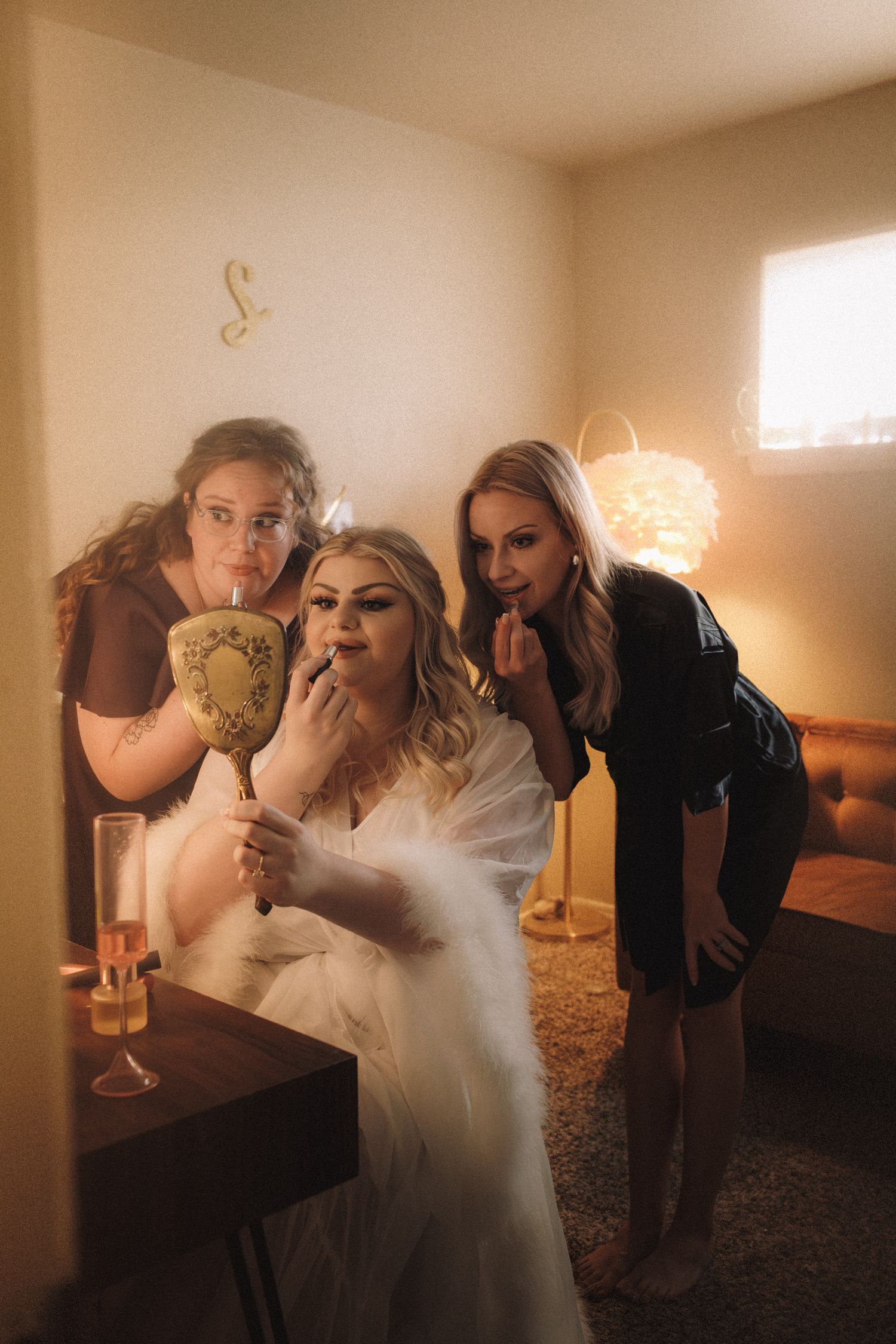 Bride in fur robe holding vintage mirror with two bridesmaids applying lipstick in warm room.