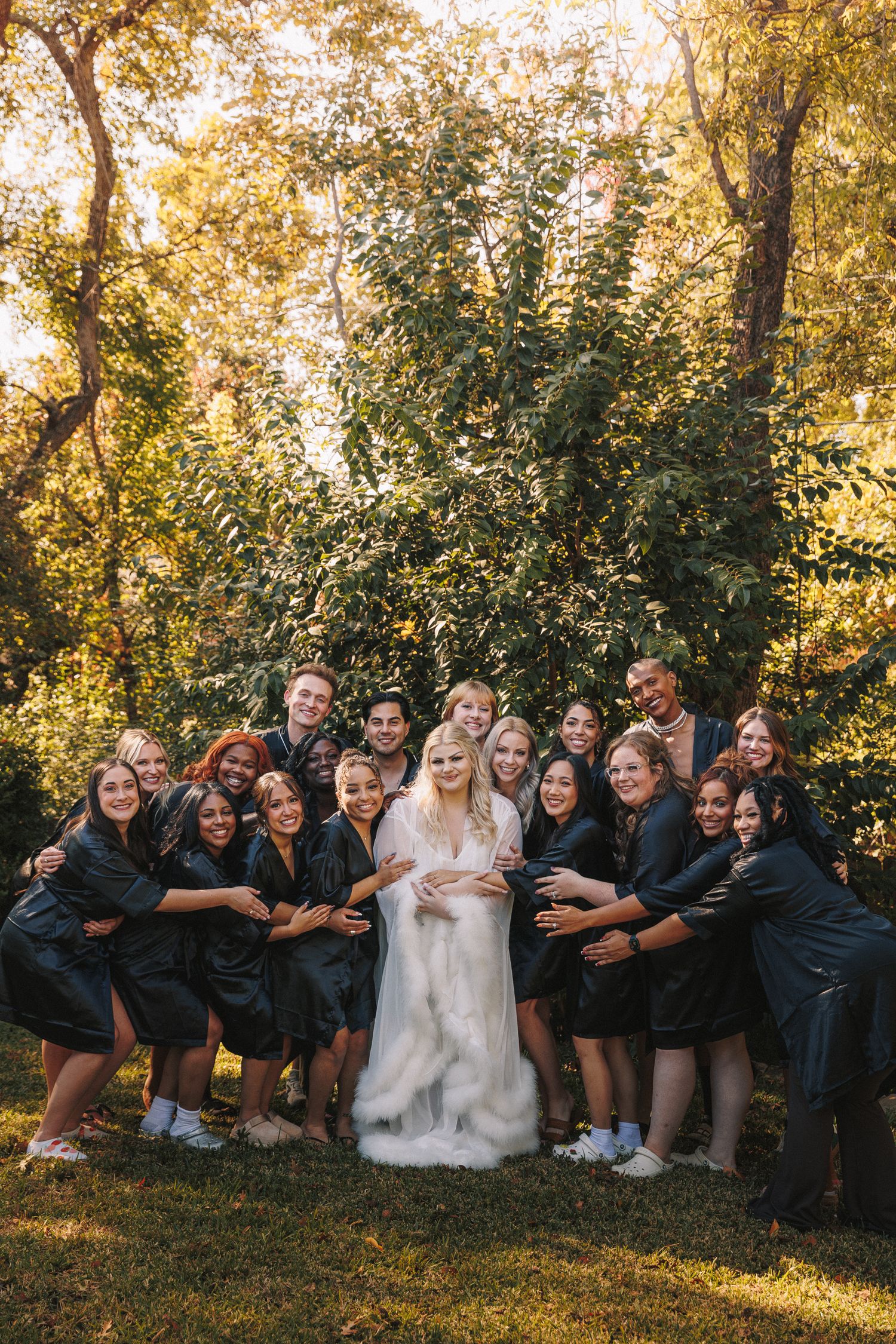 Bride in white robe surrounded by bridesmaids in black robes posing outdoors in golden autumn light.