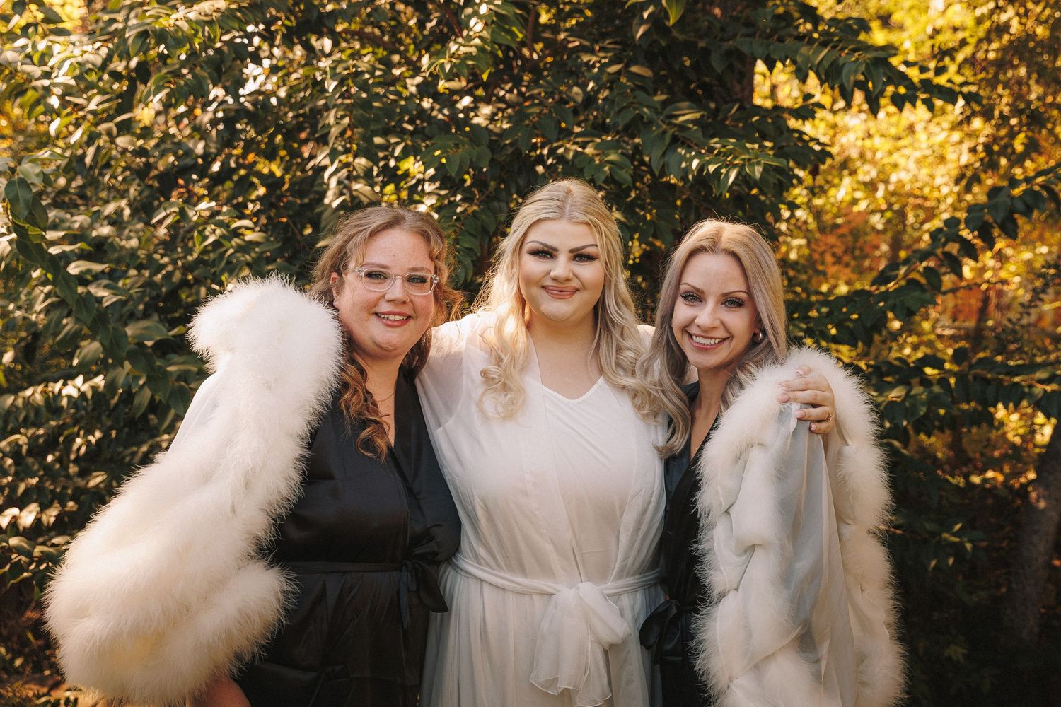 Bride and two bridesmaids in white and black robes with fur trim posing outdoors.