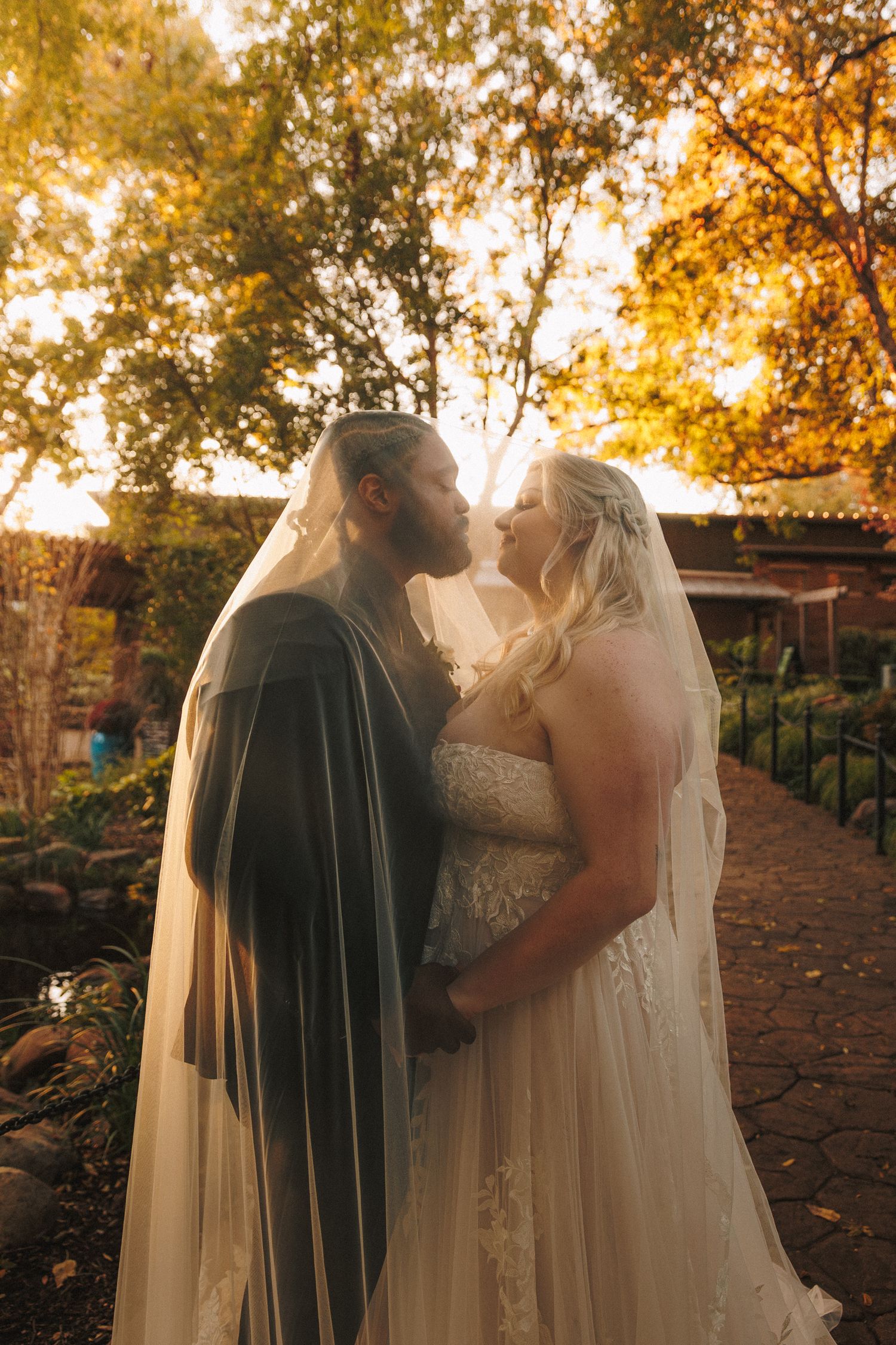 Bride and groom kissing under a shared veil in a golden sunset garden setting on their wedding day.