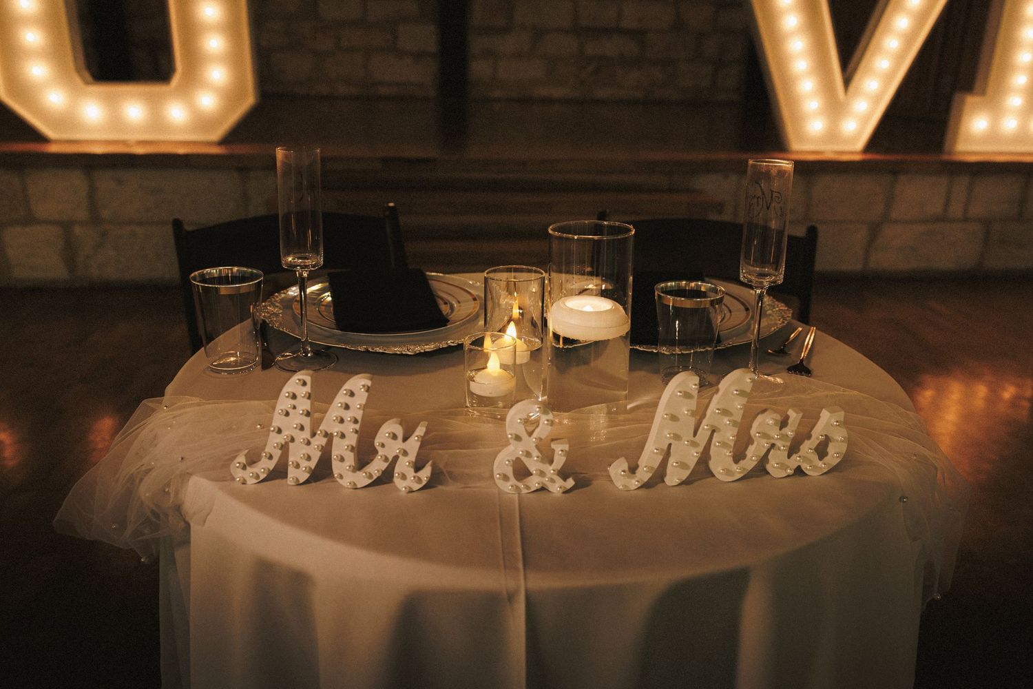 Elegant wedding sweetheart table with Mr & Mrs signs, candles, and glowing LOVE marquee letters in romantic venue.