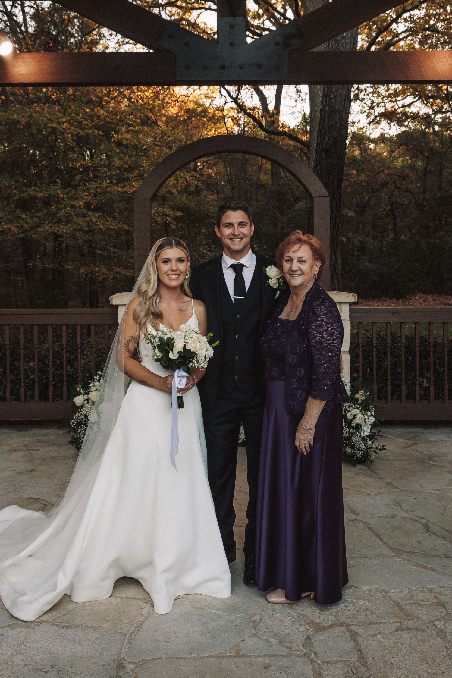 Bride in white gown and groom in black suit pose with older woman in purple lace dress at wedding venue.