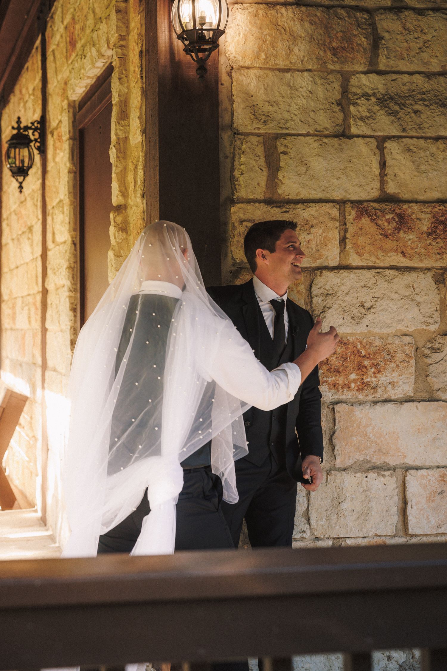 Groom smiling as bride in veil approaches for first look at stone building entrance.