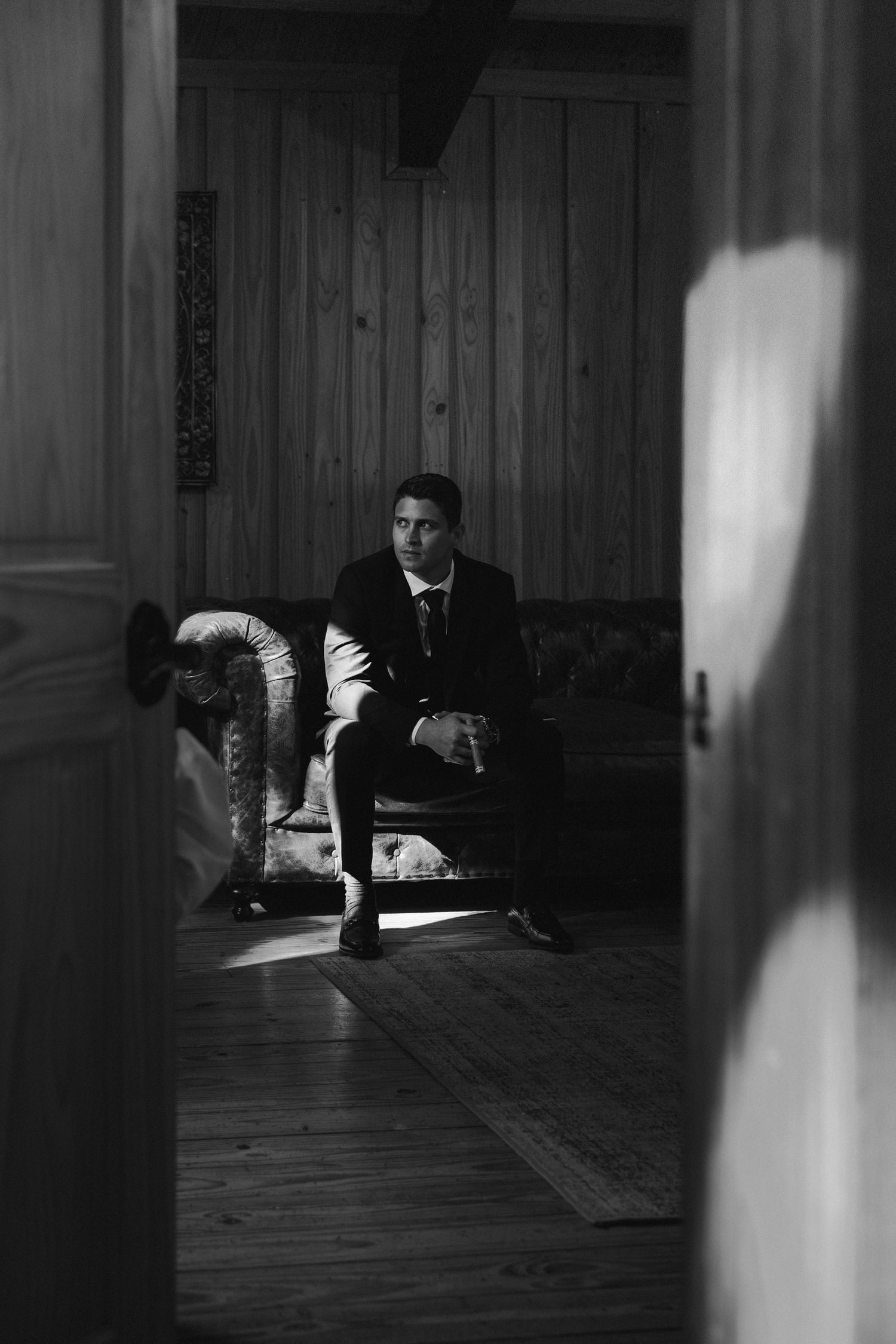 Elegant groom in black suit sitting in a leather chair in a black and white dramatic portrait.