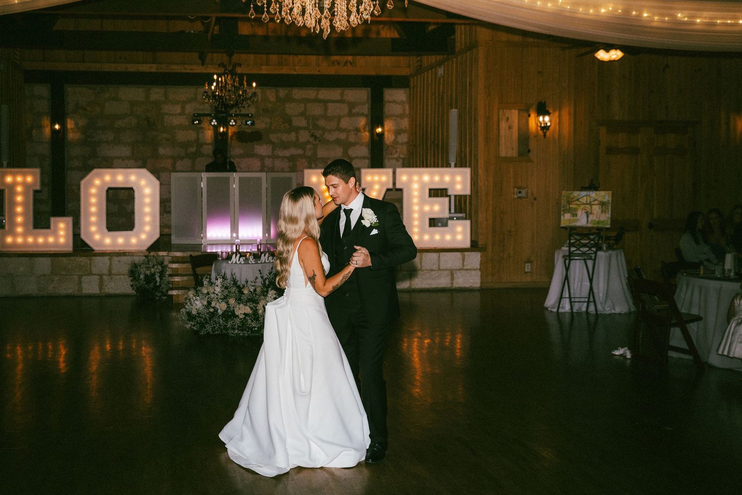 A bride and groom share their first dance in front of glowing LOVE marquee letters.
