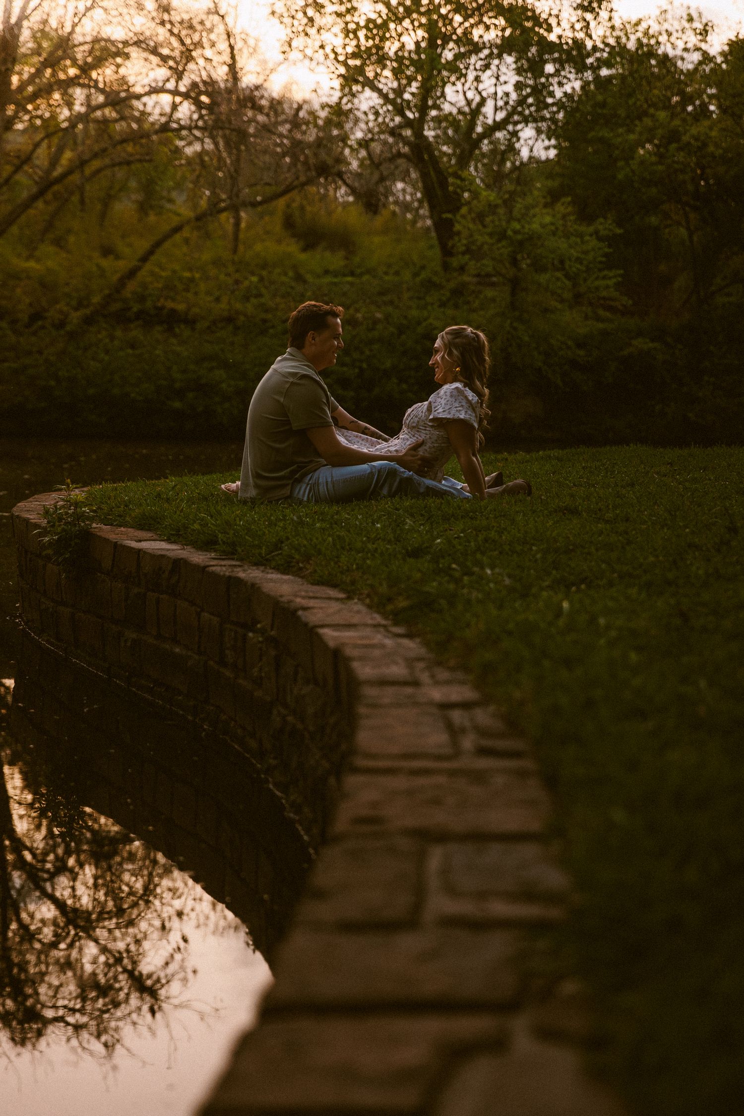 Couple sitting together on grassy bank by brick wall at sunset, surrounded by lush green trees in a romantic park setting.