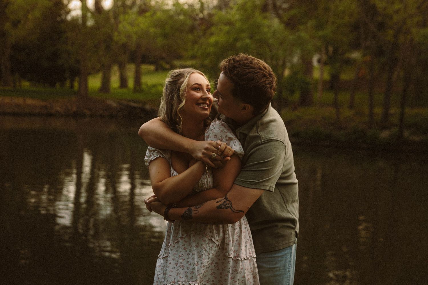 Happy couple laughing and embracing by a tranquil pond surrounded by lush green trees.