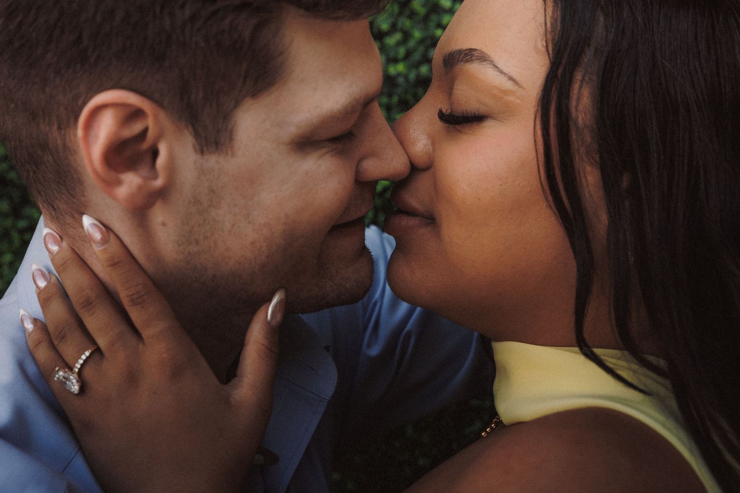 Couple sharing a romantic kiss outdoors, woman wearing an engagement ring, in a tender close-up moment.