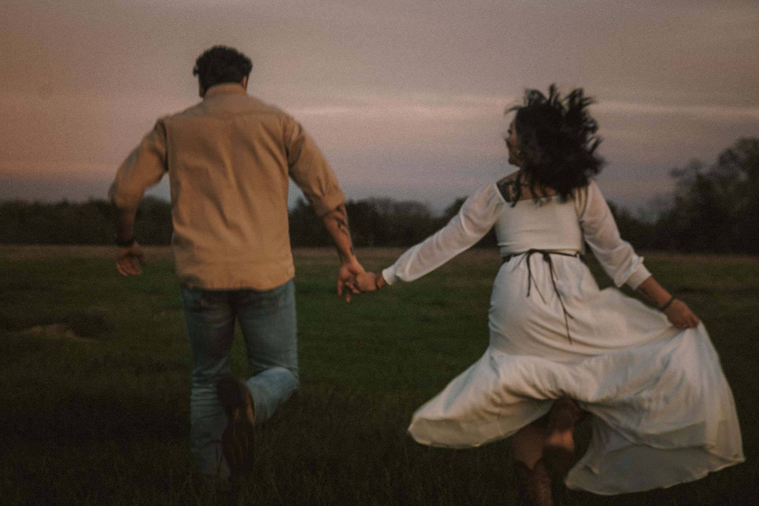 Couple running hand in hand through a green field at dusk, woman in white dress, man in beige shirt.