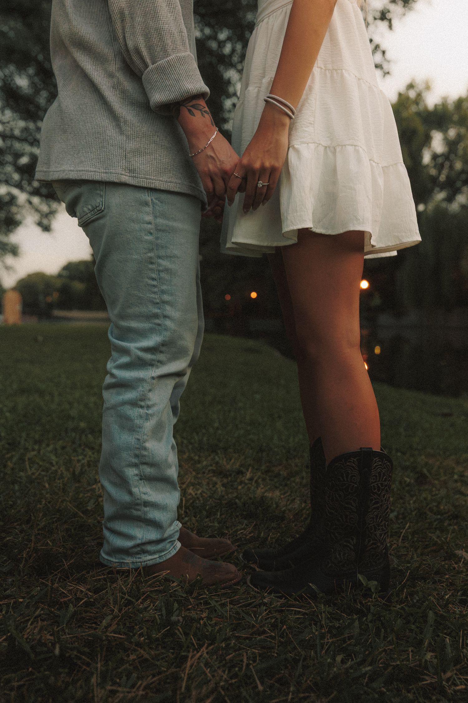 A romantic couple stands close together in a grassy field at dusk, wearing contrasting casual outfits of jeans and a dress.