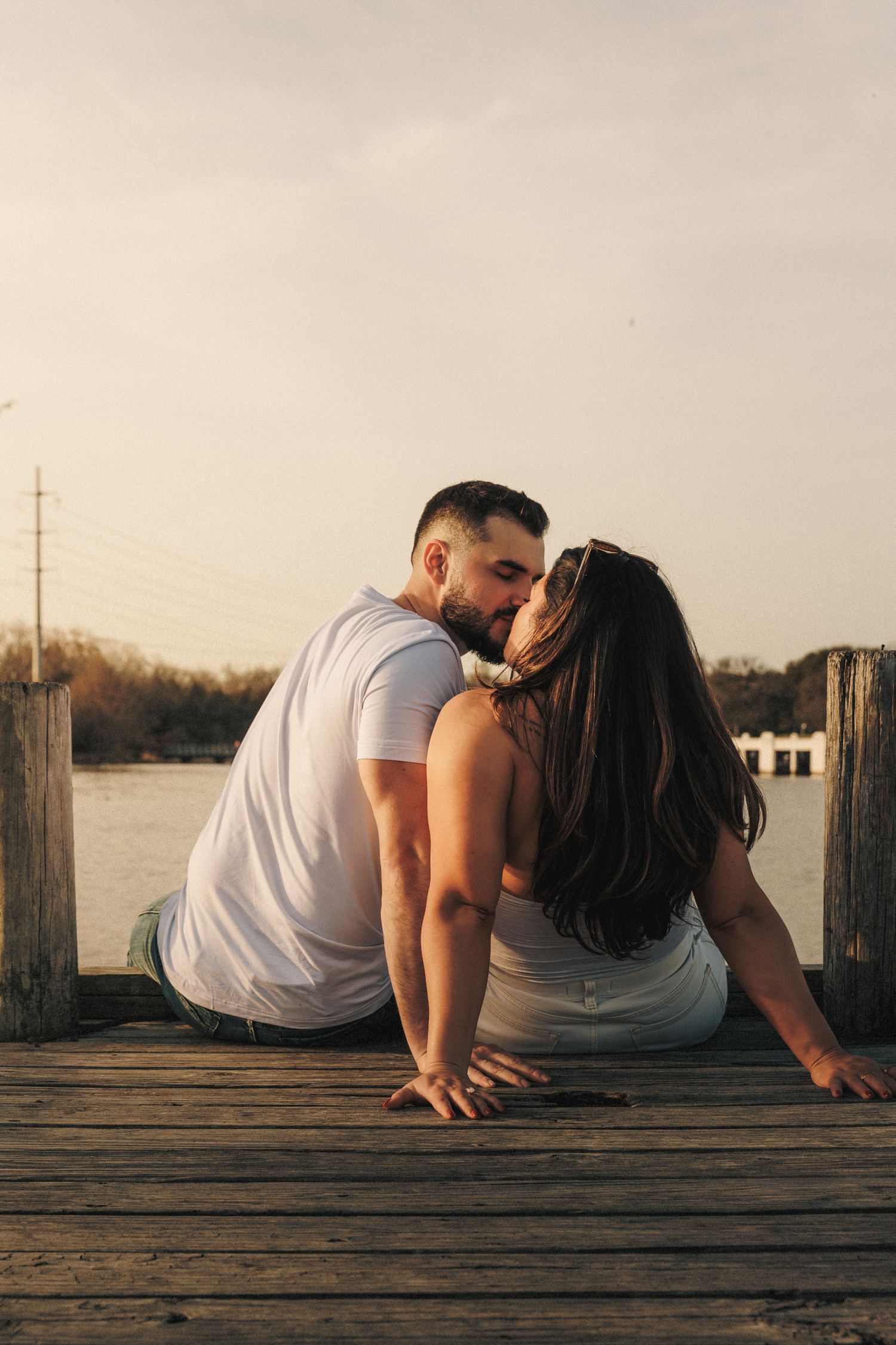 Couple relaxes on wooden dock during intimate waterfront photo session at sunset.