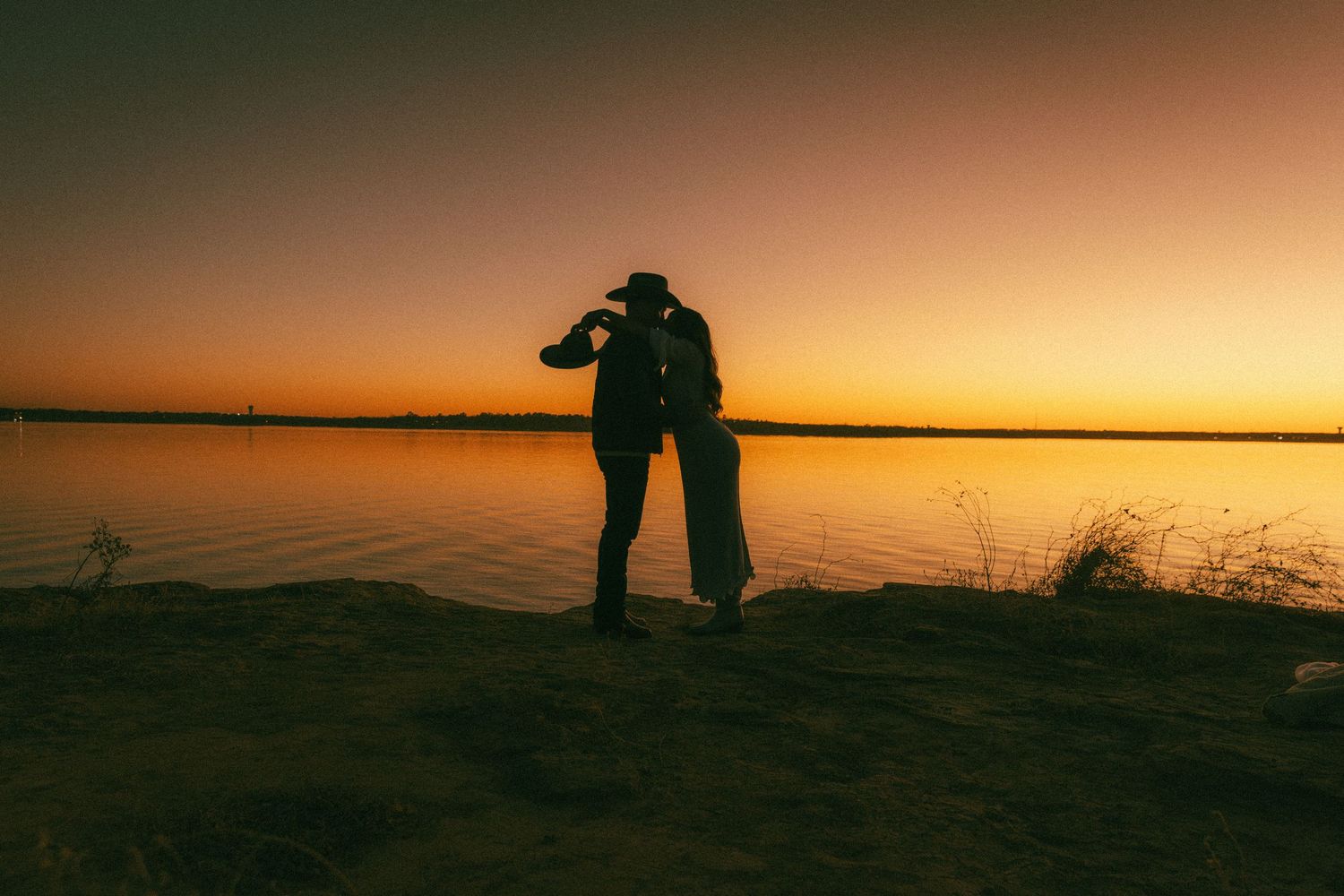 Dallas, Texas Engagement PhotograSilhouetted figures embrace against a vibrant orange sunset over a calm lake with reeds in the foreground.