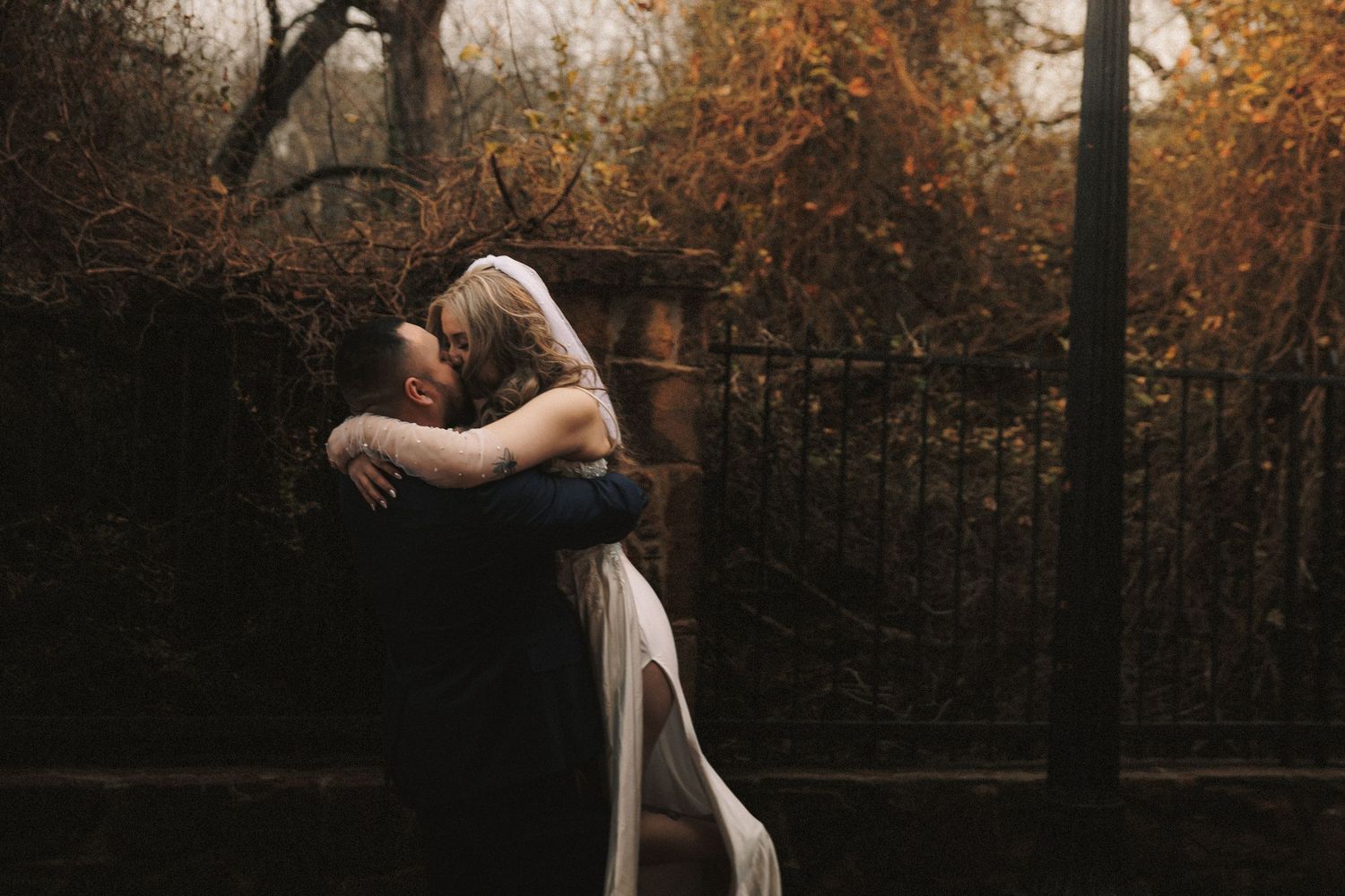 Two figures embrace in an intimate dance pose against a moody autumn backdrop with iron fencing and fallen leaves.