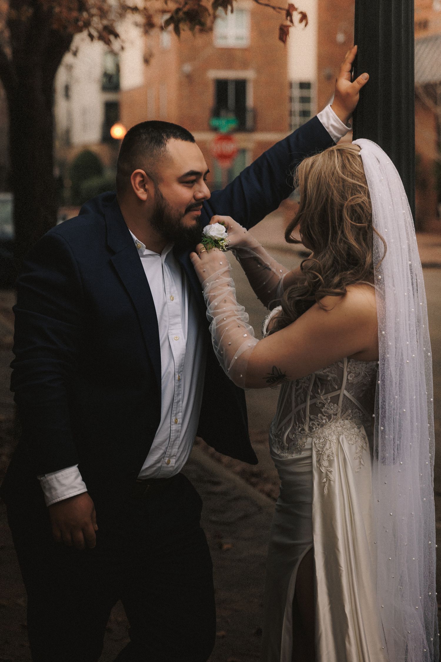 A couple shares an intimate dance moment outdoors with brick buildings and autumn trees in the background.