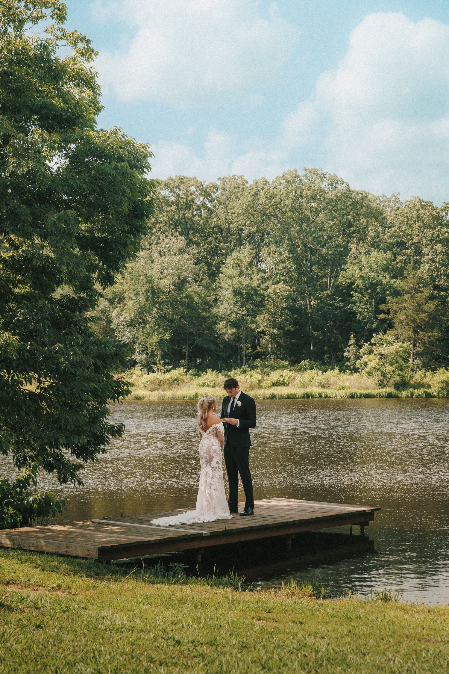 Couple in wedding attire stands on wooden dock overlooking serene lake surrounded by lush green trees.