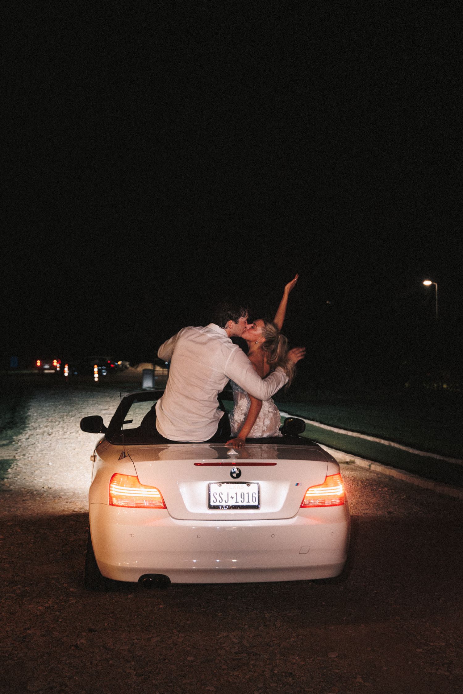 Romantic nighttime photo of newlyweds posing on the trunk of a car with taillights glowing.